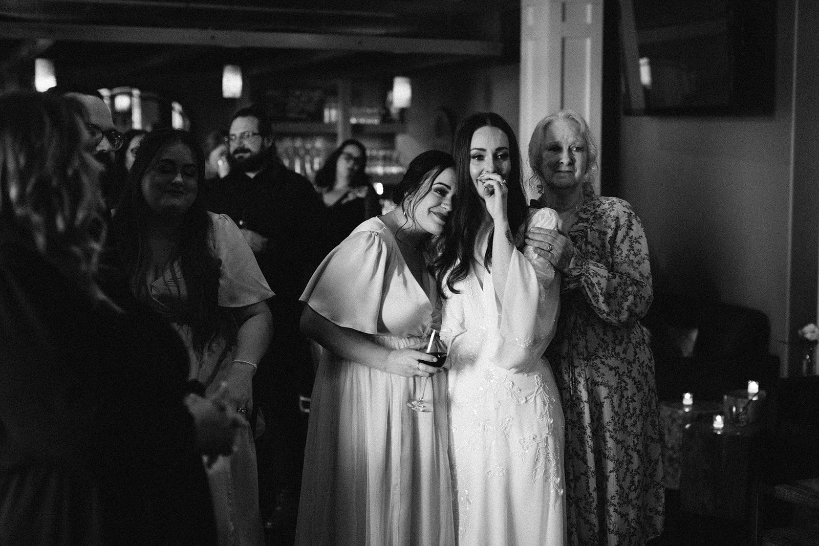 Black and white photo of a group of women, with two women in the center wearing wedding dresses, surrounded by other guests at a celebration in a cozy indoor setting.