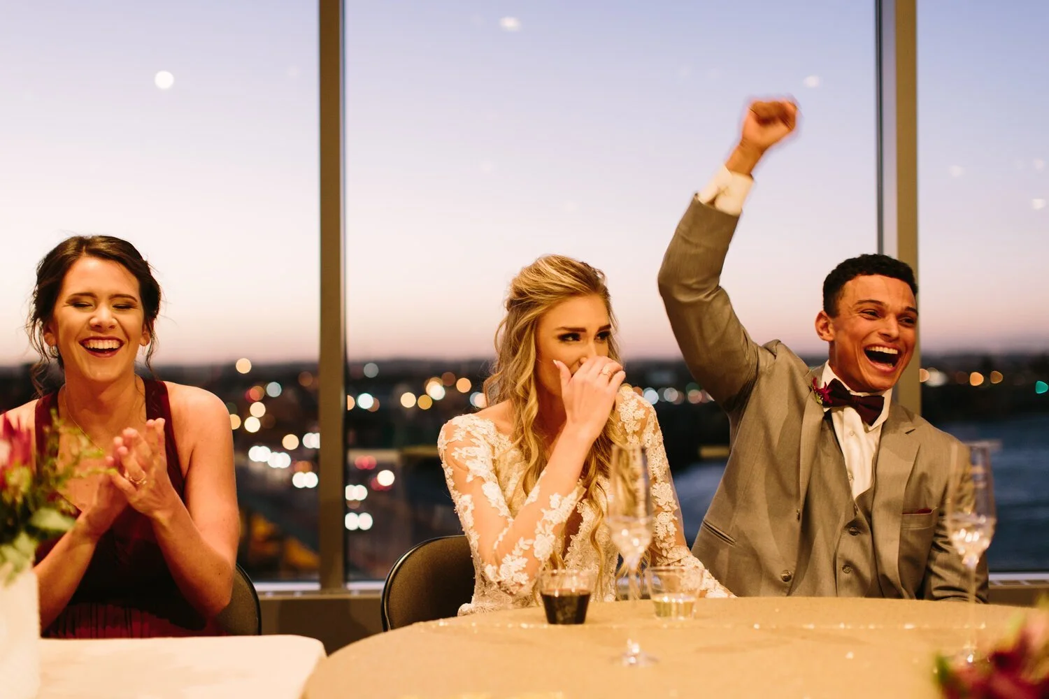 People celebrating at a wedding reception with a view of a city skyline in the background; the groom is raising his arm in excitement, the bride is covering her mouth while laughing, and a woman on the left is smiling and clapping.