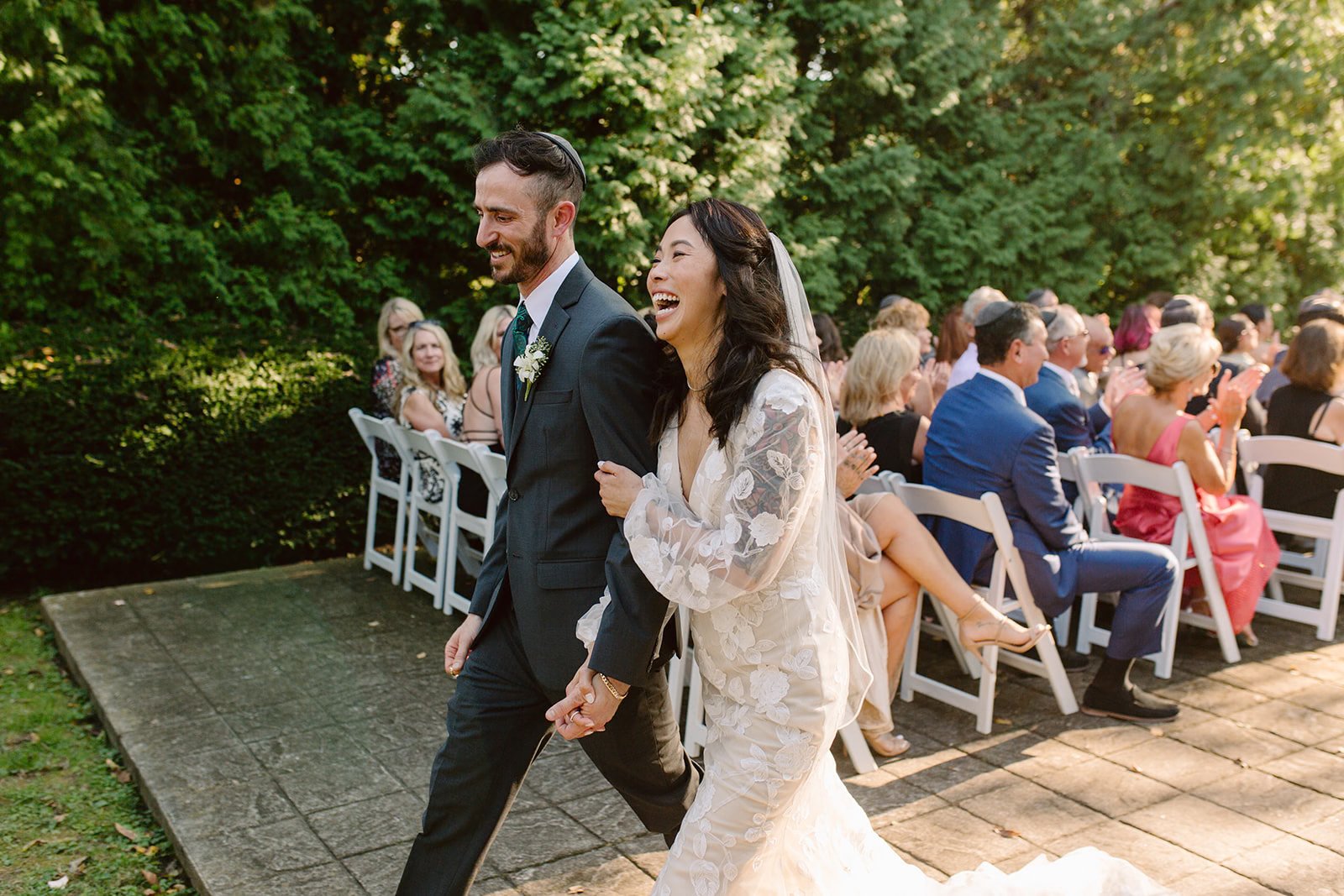 Bride and groom walking down the aisle at their outdoor wedding, smiling and holding hands, with seated guests clapping in the background.