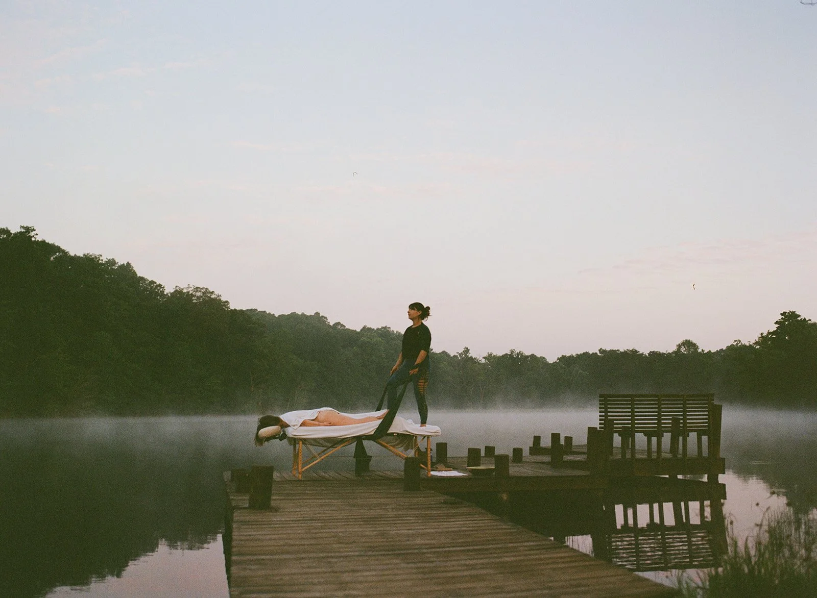 A woman lies peacefully on a massage table on a wooden dock by a misty lake during dawn, as another woman stands beside her, holding a strap, with trees and a cloudy sky in the background.