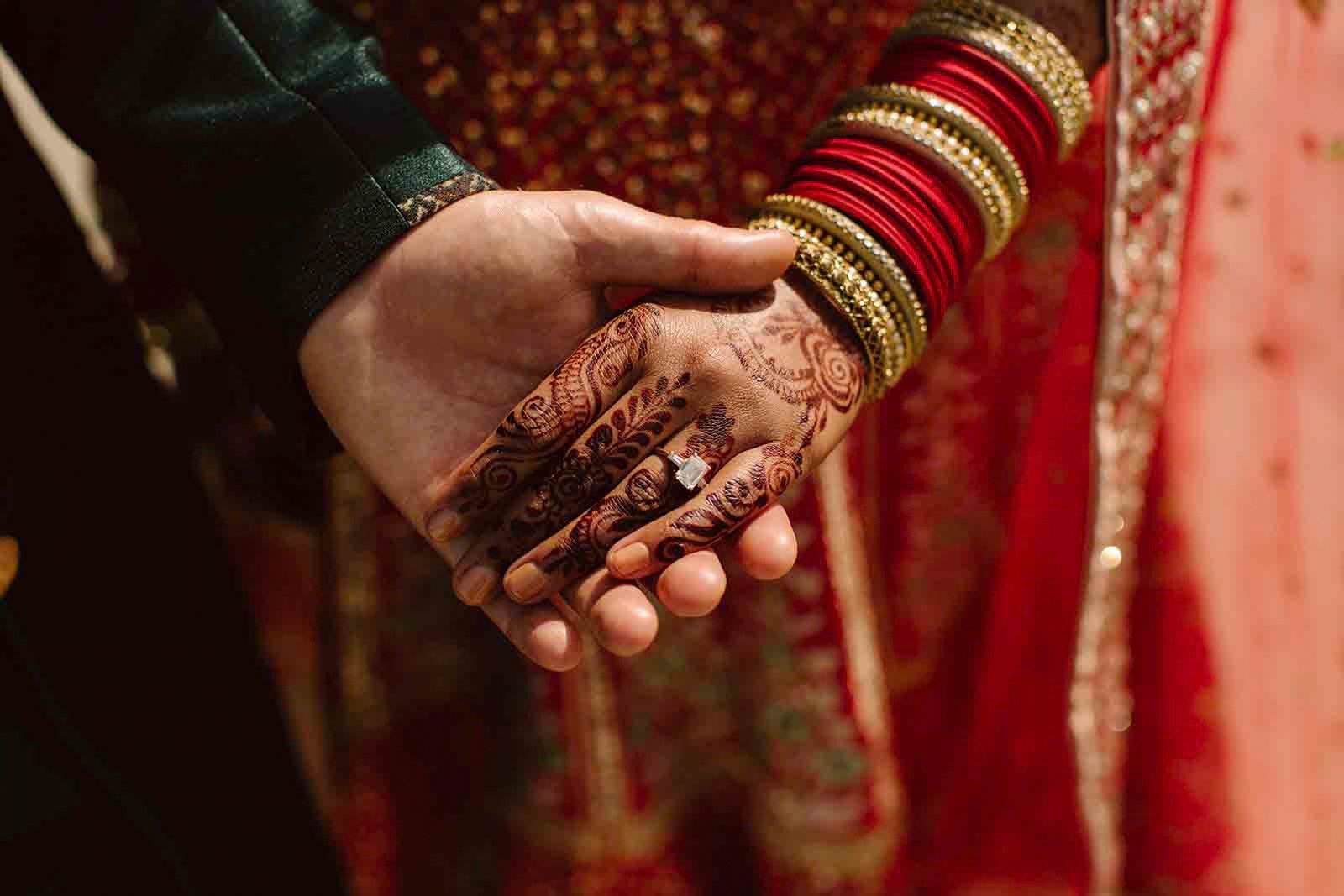 Close-up of a bride and groom holding hands during a wedding ceremony. The bride's hand features intricate henna designs and a prominent ring, while her wrist is adorned with multiple red and gold bangles. The groom's hand is gently holding hers against a traditional red and gold wedding attire.