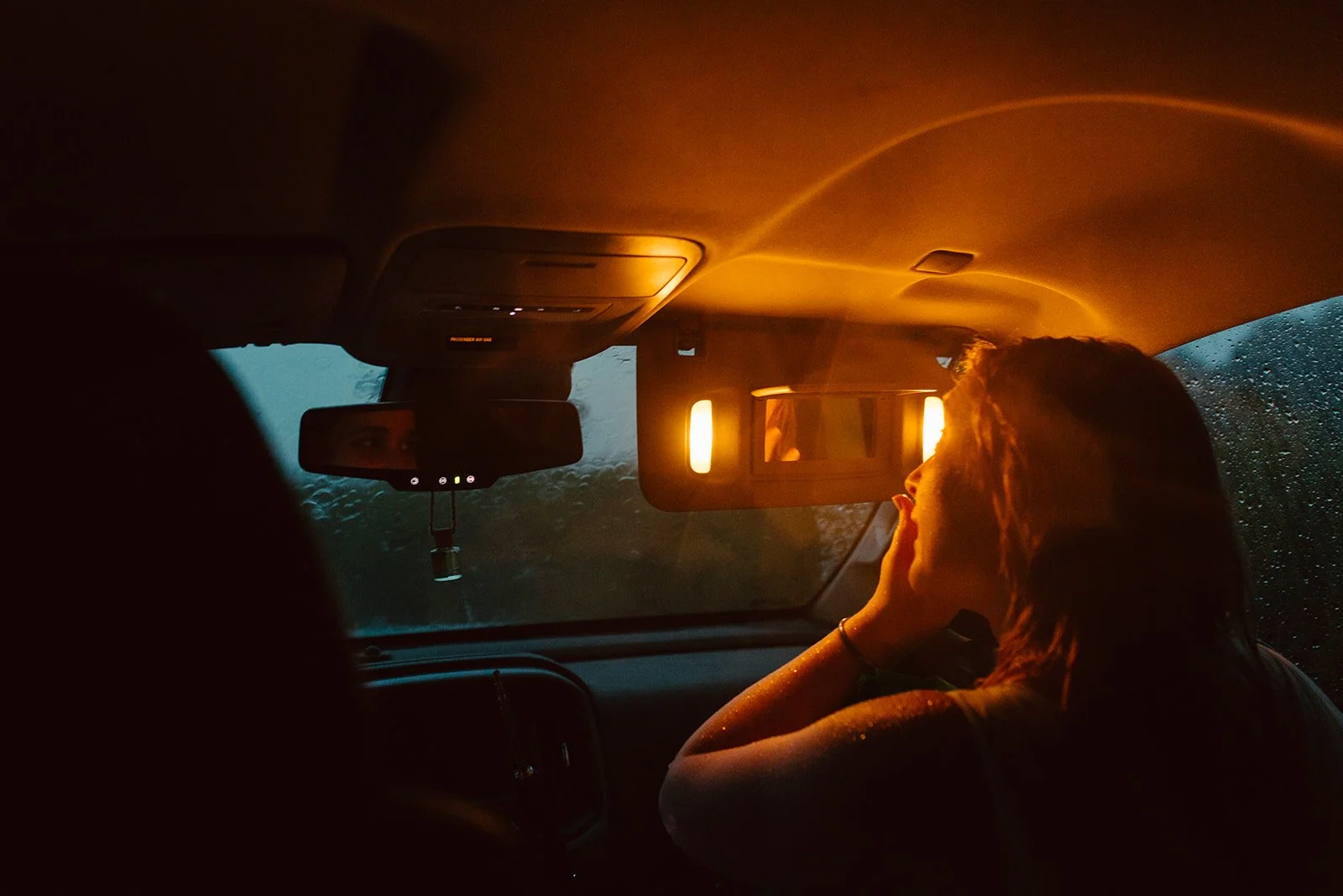 A woman sitting in a car during rain at sunset, looking out the window.
