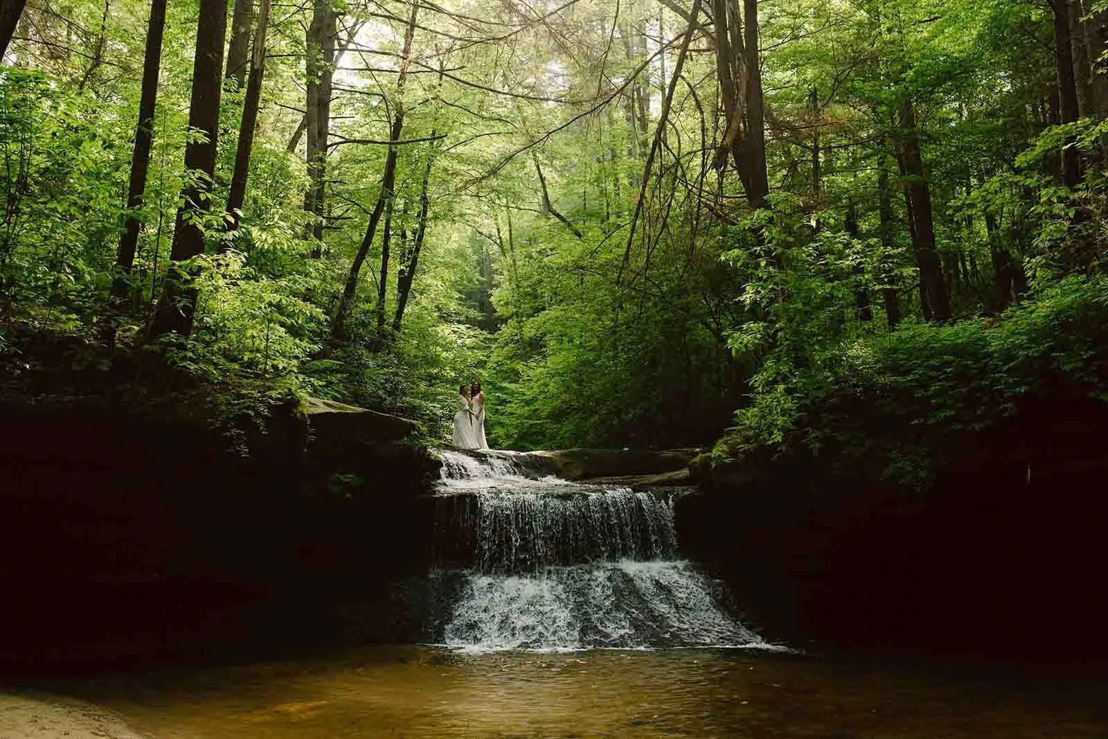 Two women in white dresses standing on rocks beside a small waterfall in a lush, green forest.