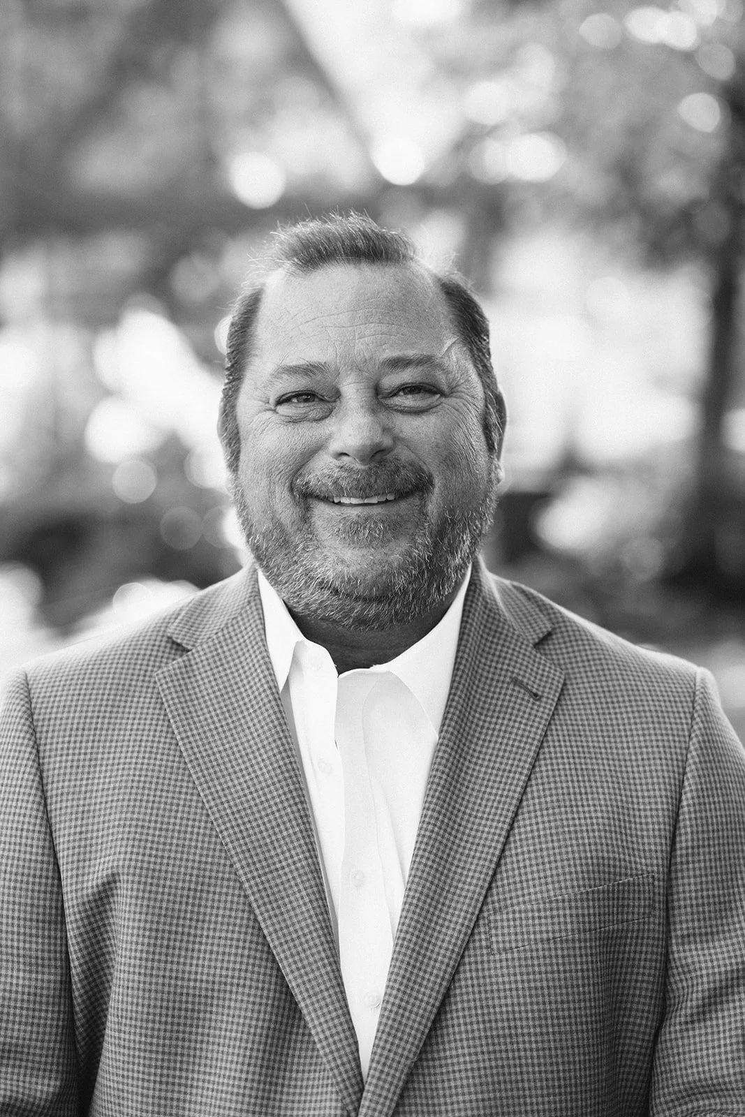 A black and white portrait of a smiling man wearing a blazer and a white collared shirt, outdoors with blurred trees and sunlight in the background.