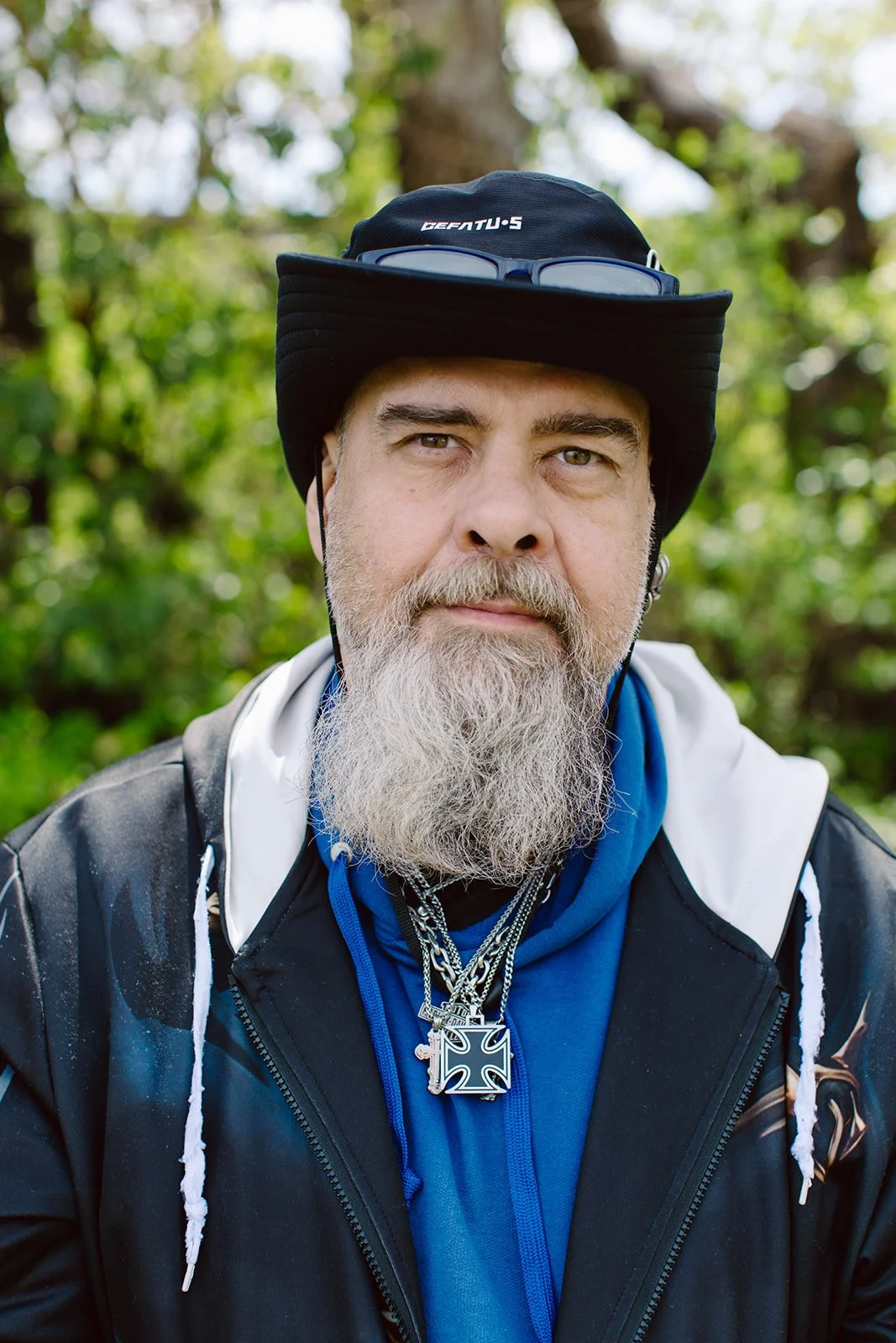 A middle-aged man with a gray beard and mustache, wearing a black hat, sunglasses resting on the hat, layered necklaces, a black jacket over a blue hoodie, standing outdoors with a green, wooded background.