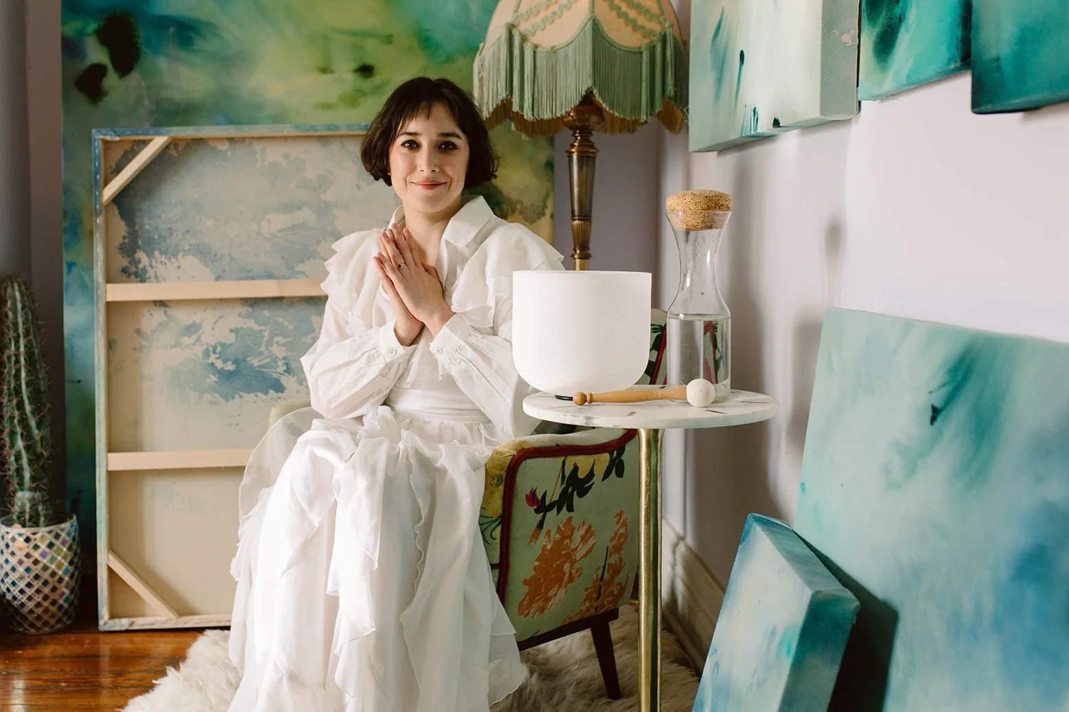 A woman sitting on a patterned chair with a white dress, hands folded on her chest, in an art studio with abstract paintings, a lamp, and art supplies around.