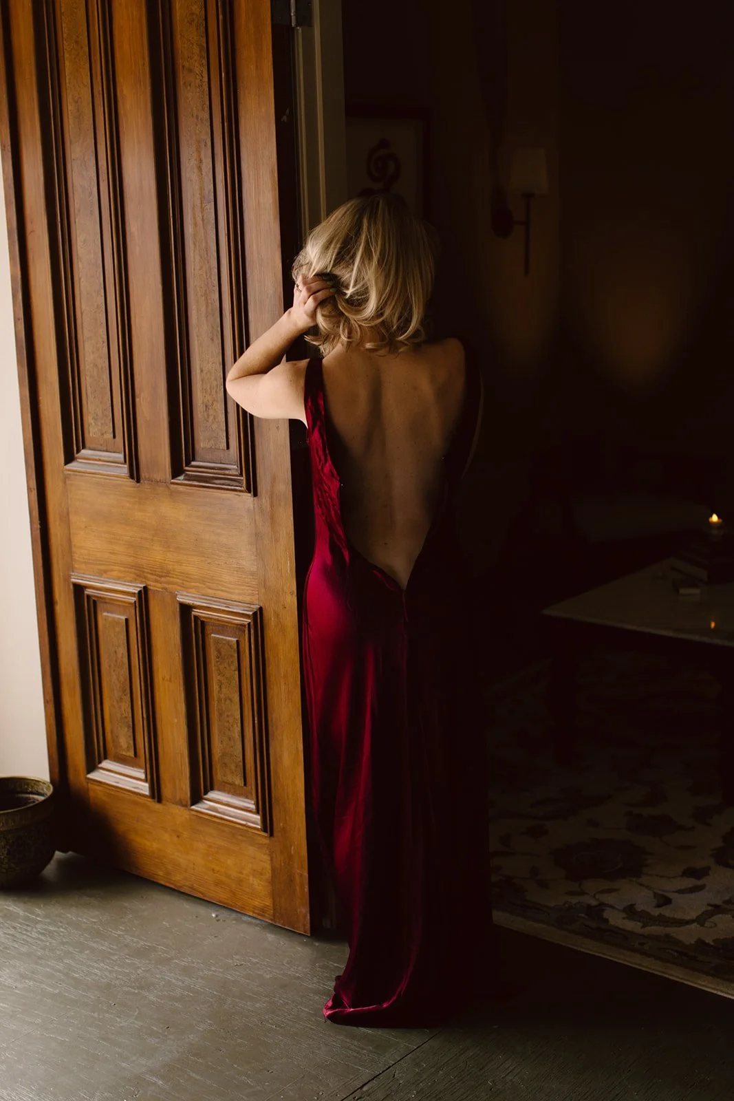 A woman with blonde hair in a red satin dress stands in a doorway, facing away, with her hand in her hair.