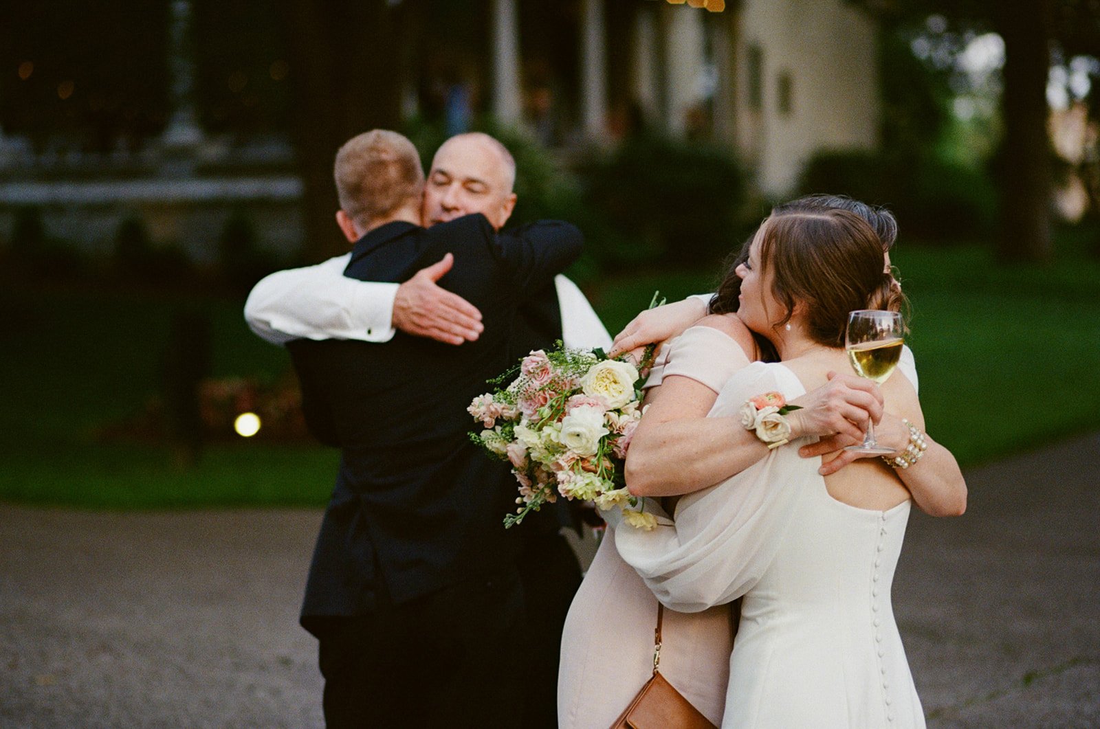 Two women hugging and embracing at a wedding reception, holding wine glasses, with flowers. Two men hugging in the background.