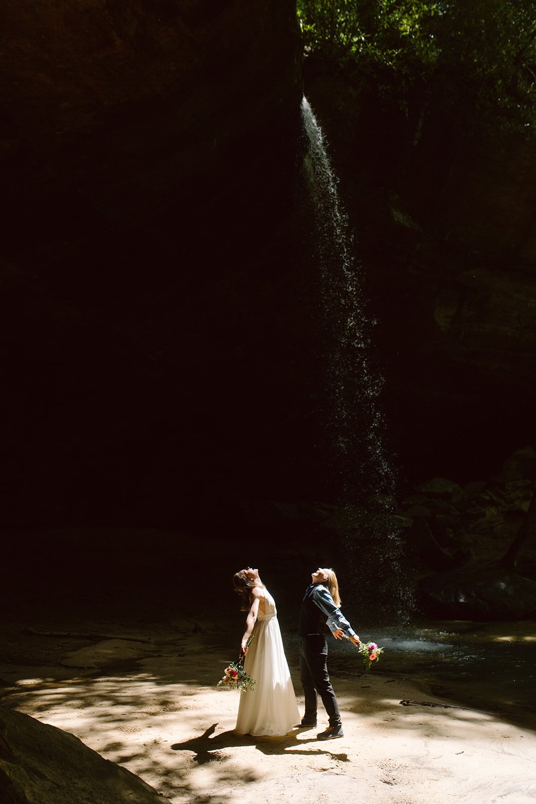 Two women, one in a wedding dress and the other in casual clothing, holding bouquets, standing under a waterfall in a dark forested area, with sunlight filtering through the trees.