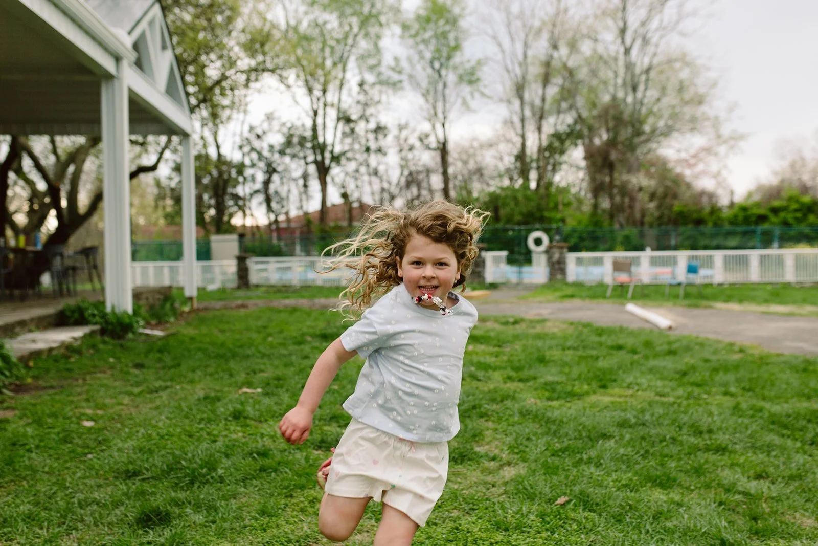 A young girl with curly hair running on a grassy backyard, smiling while her hair blows back.