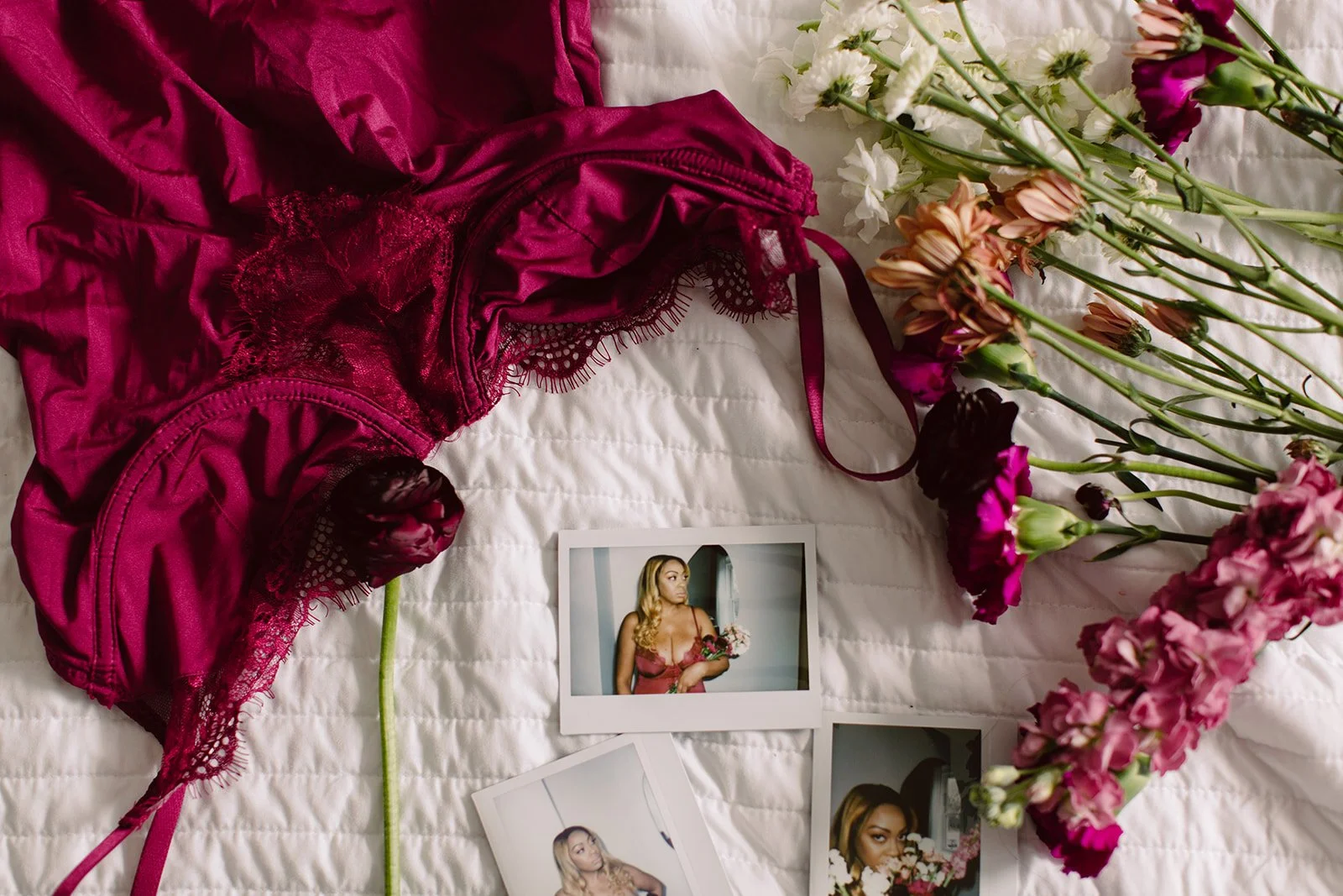 Pink lace lingerie, a bouquet of mixed pink and white flowers, and three instant photographs of a woman holding flowers on a white quilted bedspread.