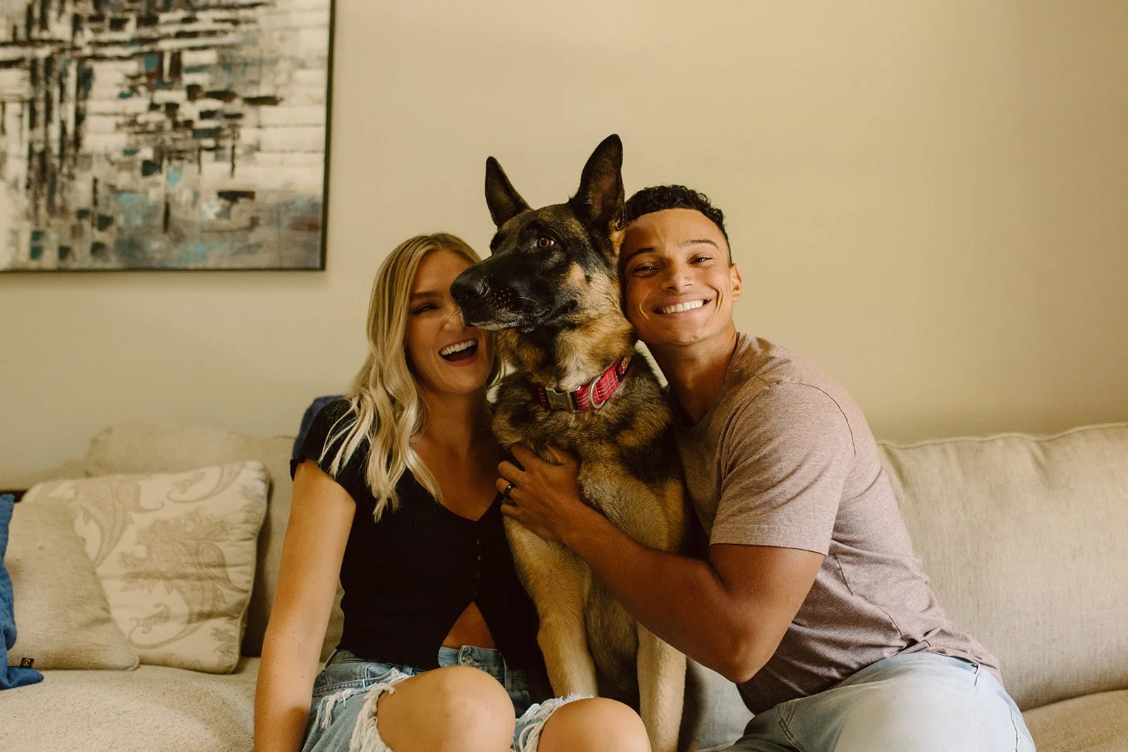 A happy couple sitting on a beige sofa, holding a large German Shepherd dog between them. The woman has long blonde hair and is smiling, wearing a black top and denim shorts. The man has short dark hair, smiling broadly, wearing a light brown T-shirt and light-colored pants. They are inside a living room with a beige wall and a large abstract painting hanging behind them.
