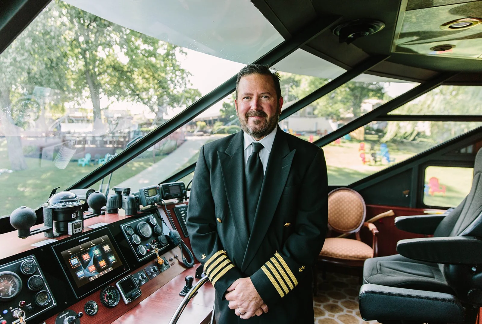 A man in a black captain's uniform with gold stripes on the sleeves standing inside the bridge of a boat, with a view of a park outside.