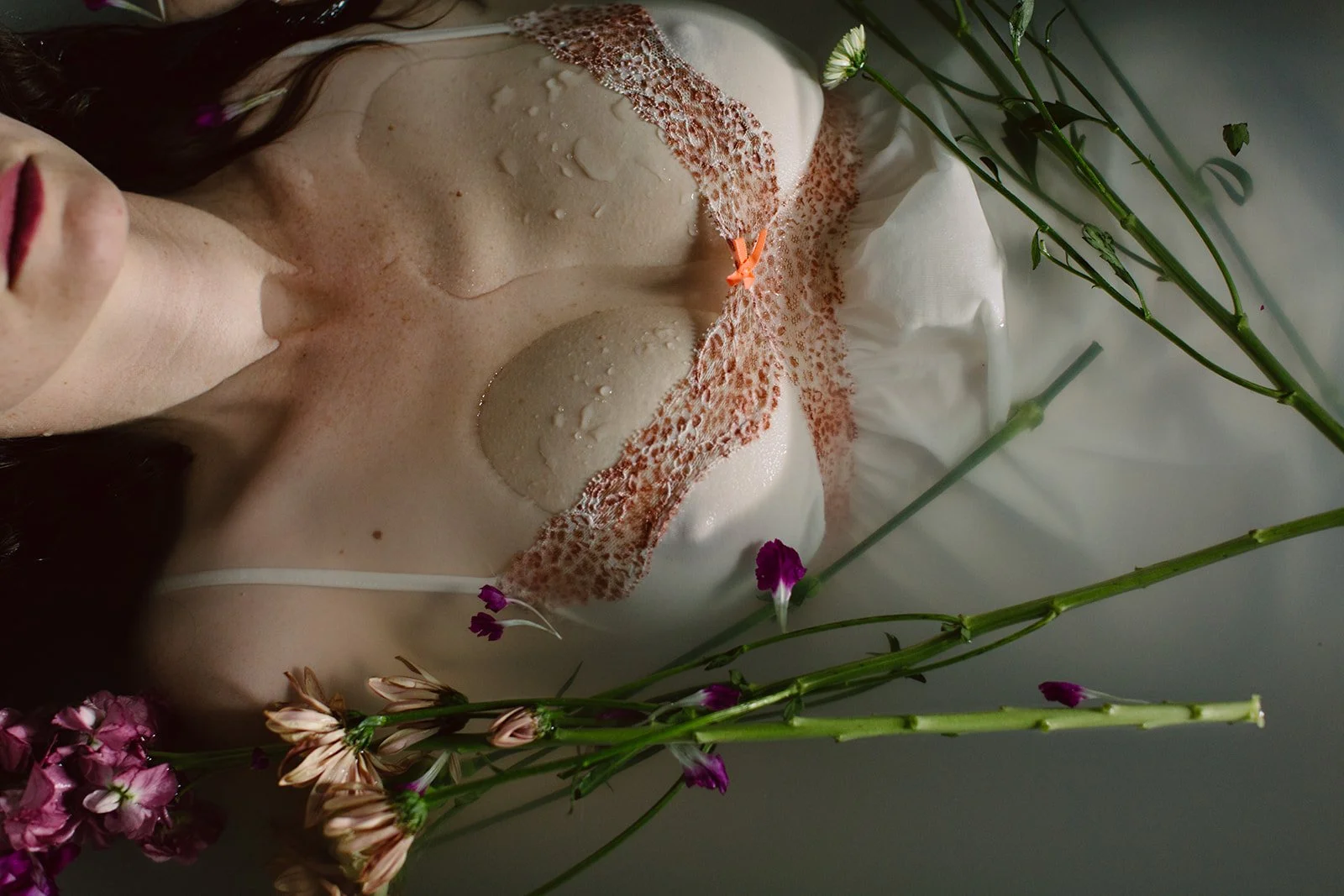 Close-up of a woman with wet skin and flowers around her chest and shoulders, lying in water.