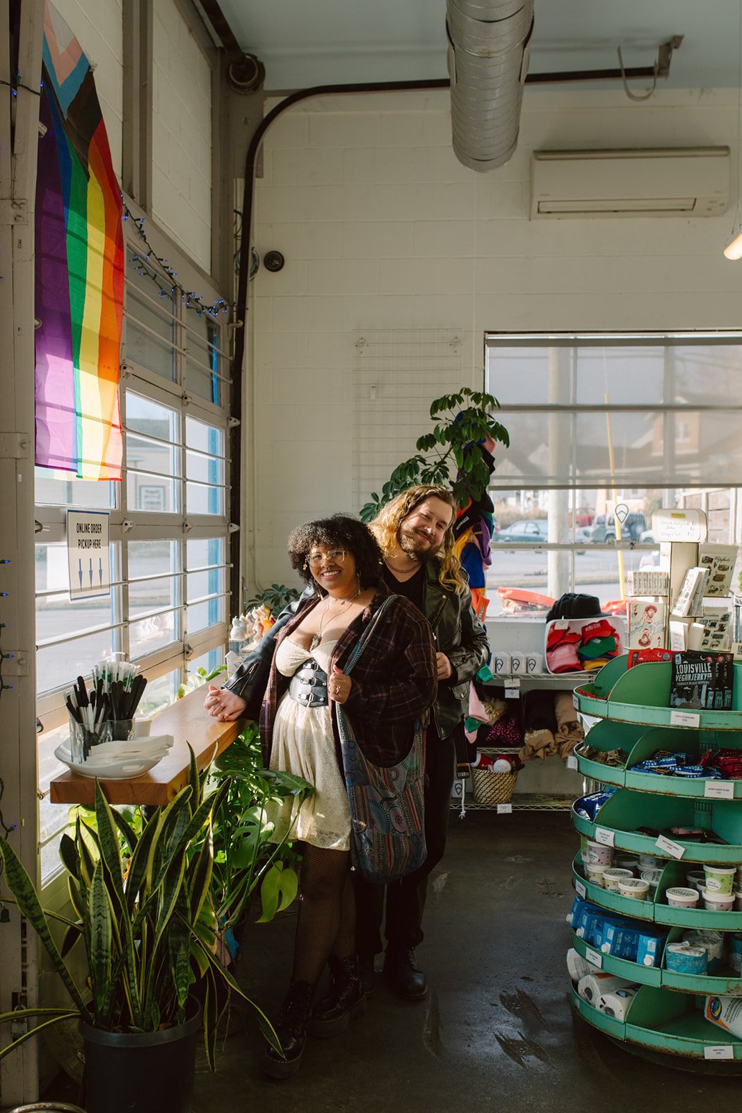 Two smiling people standing inside a store near a window with a rainbow pride flag. They are surrounded by plants and store shelves with various items, including snacks and gifts.