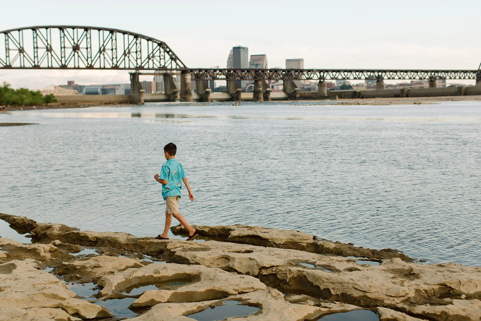 A boy walking on rocks along a river with a bridge and city skyline in the background.