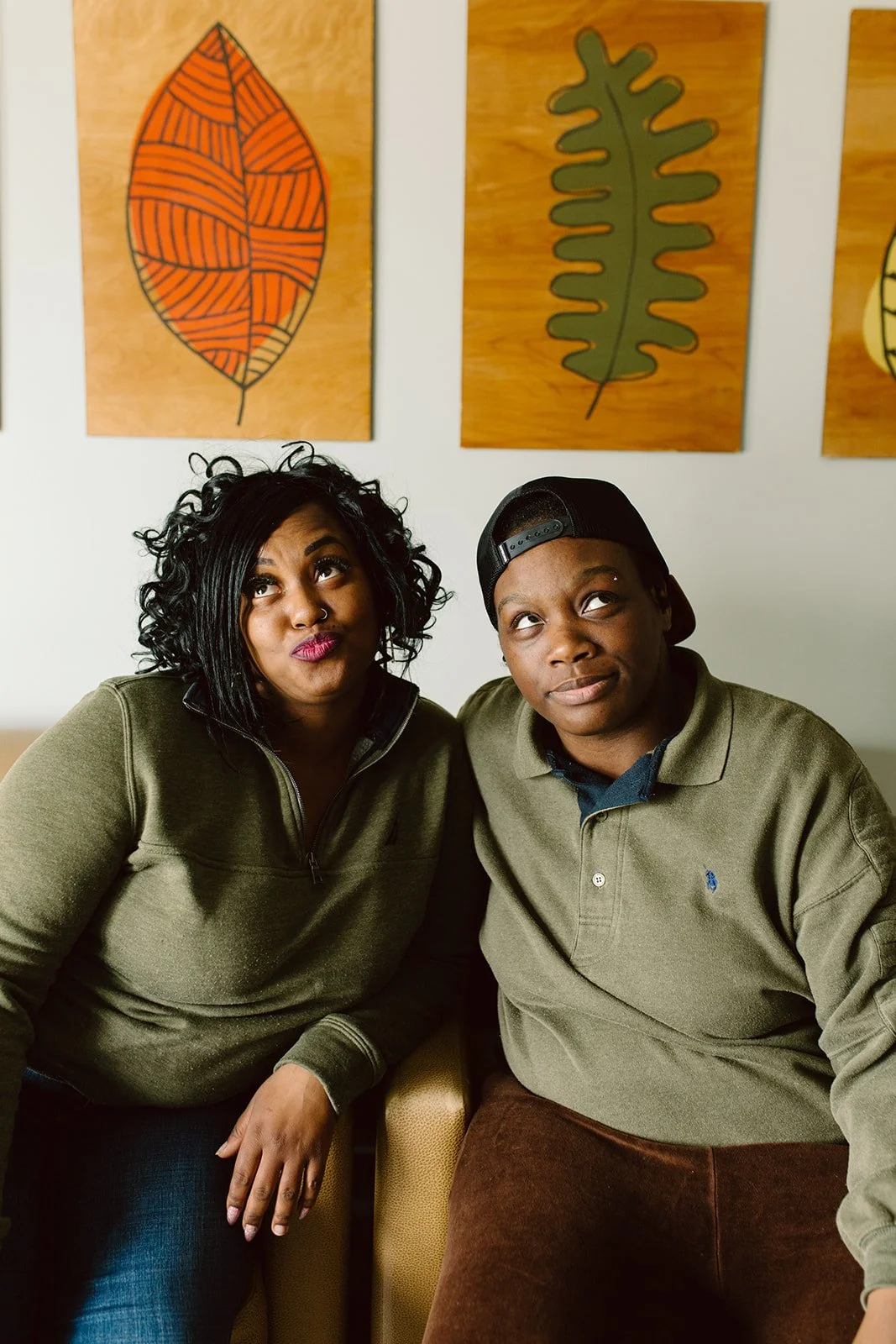 Two young people sitting close together in a casual setting, looking up and to the side, against a wall with framed art of stylized leaves.