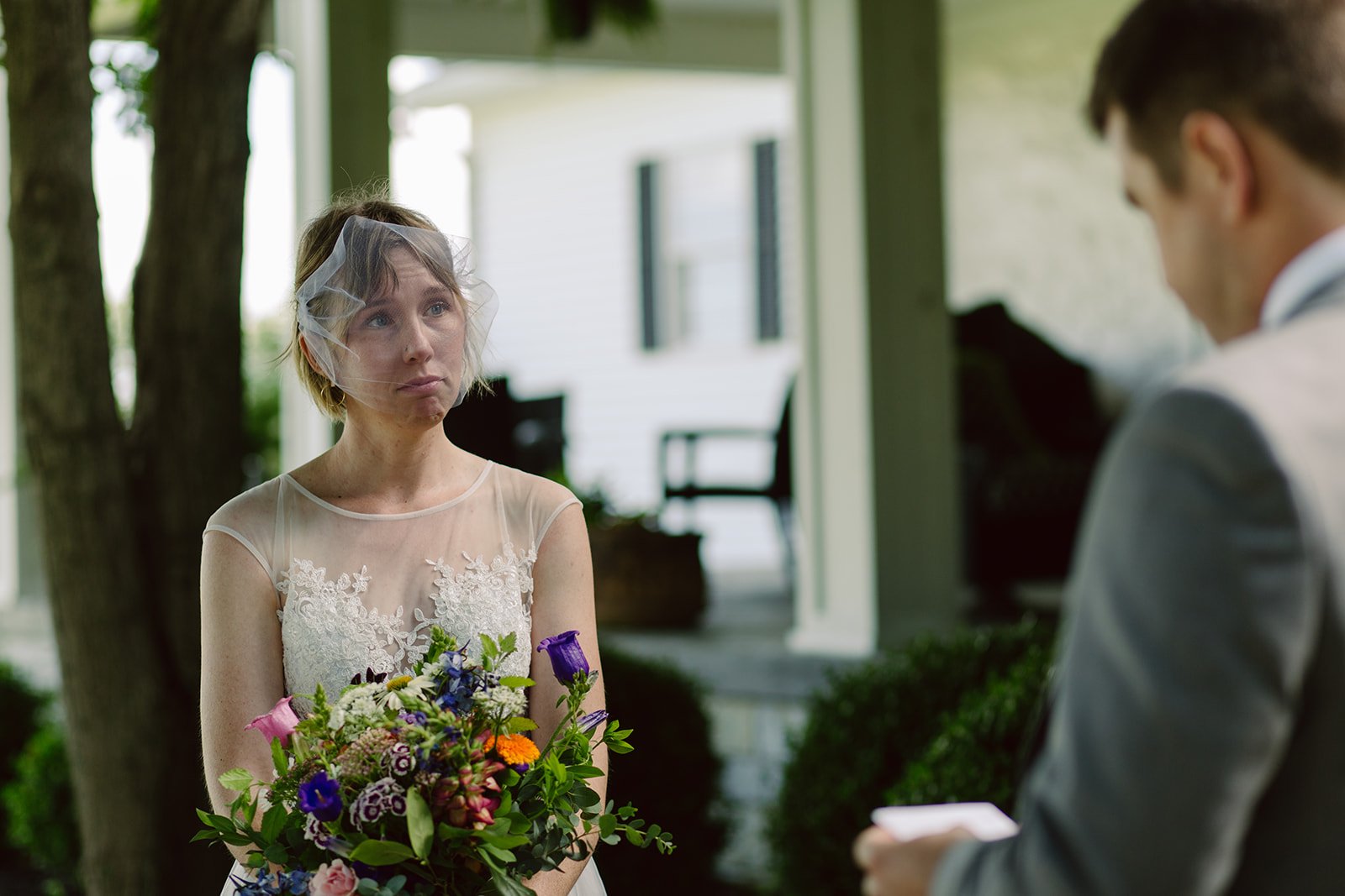 A woman in a wedding dress holding a colorful bouquet looks emotional during a wedding ceremony, with a man in a suit reading vows or a speech.