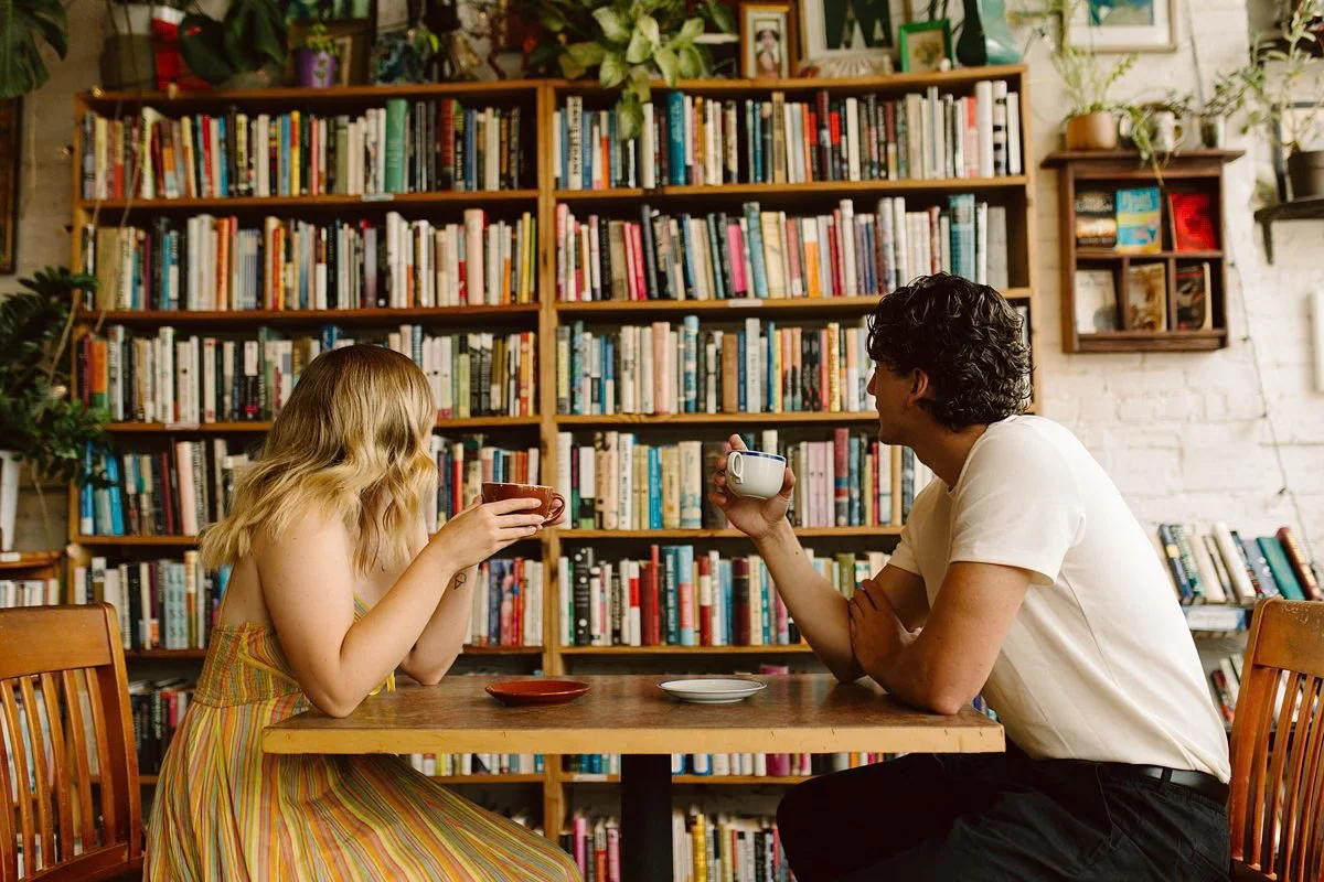 A young woman and a man sit across from each other at a wooden table, sipping coffee or tea in a cozy, book-filled space with shelves filled with colorful books and plants.