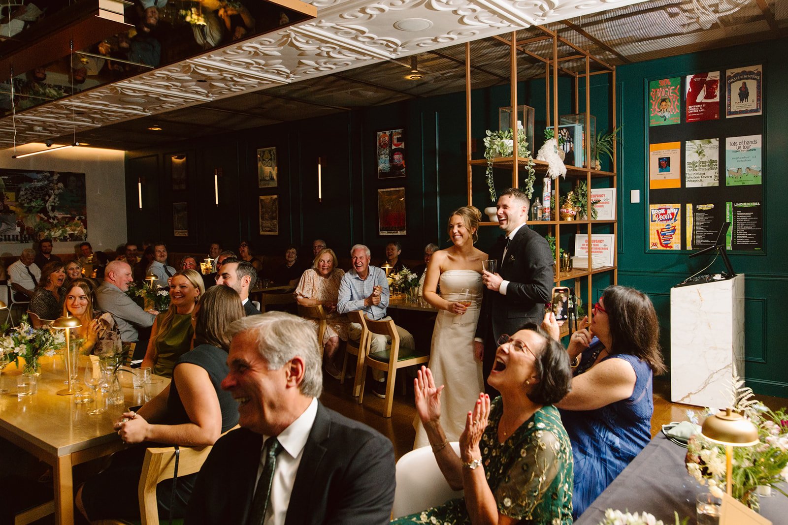 Wedding reception with bride and groom standing together, guests sitting and laughing, decorated with flowers and candles in a restaurant.