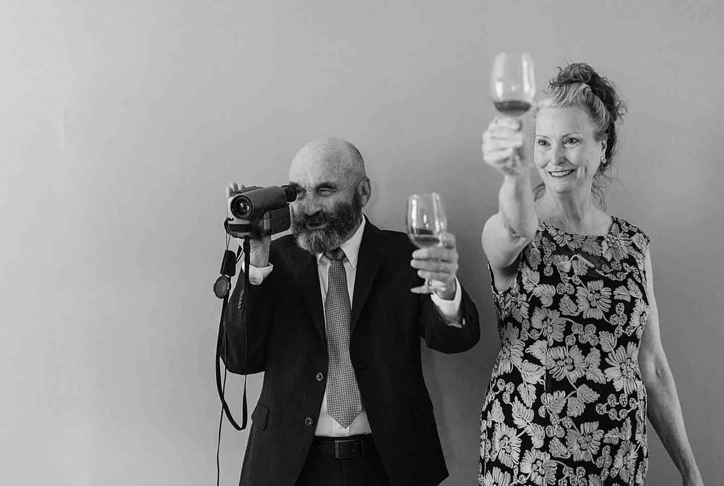 A black-and-white photo of a man with a bald head and beard wearing a suit, using a camcorder, standing next to a woman with curly hair in a floral dress, smiling, holding a glass of wine and raising it in a toast.