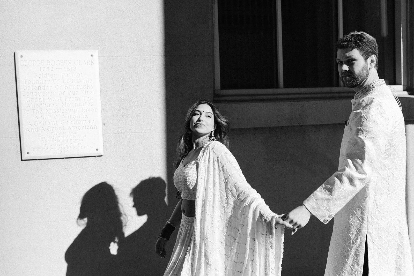 A black-and-white photo of a man and woman holding hands, standing against a wall with a plaque. The woman is smiling and wearing a traditional Indian outfit, while the man is dressed in a white sherwani. Their shadows are cast on the wall next to them.
