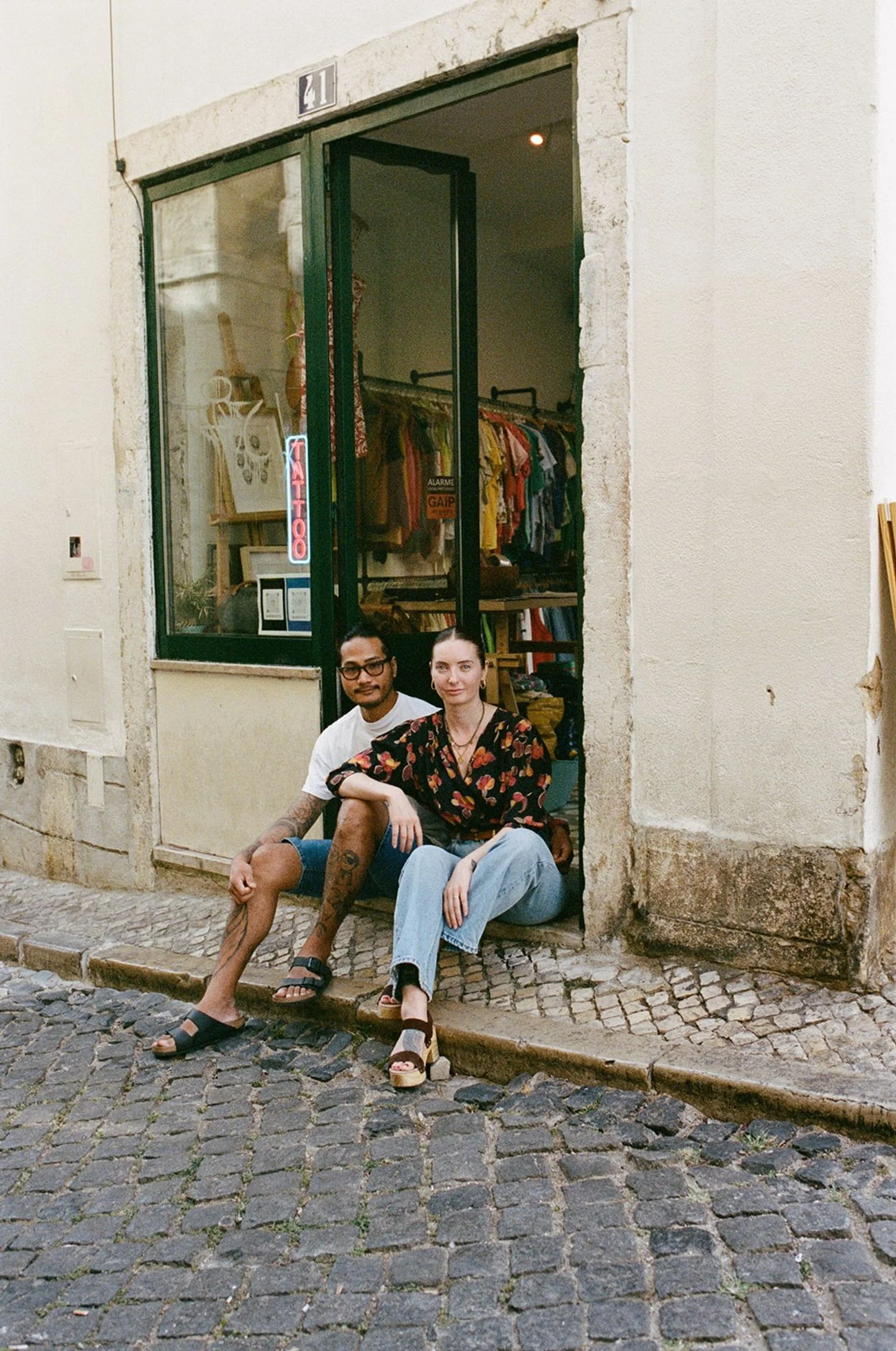 Two people sitting in front of a small clothing store on a cobblestone street.