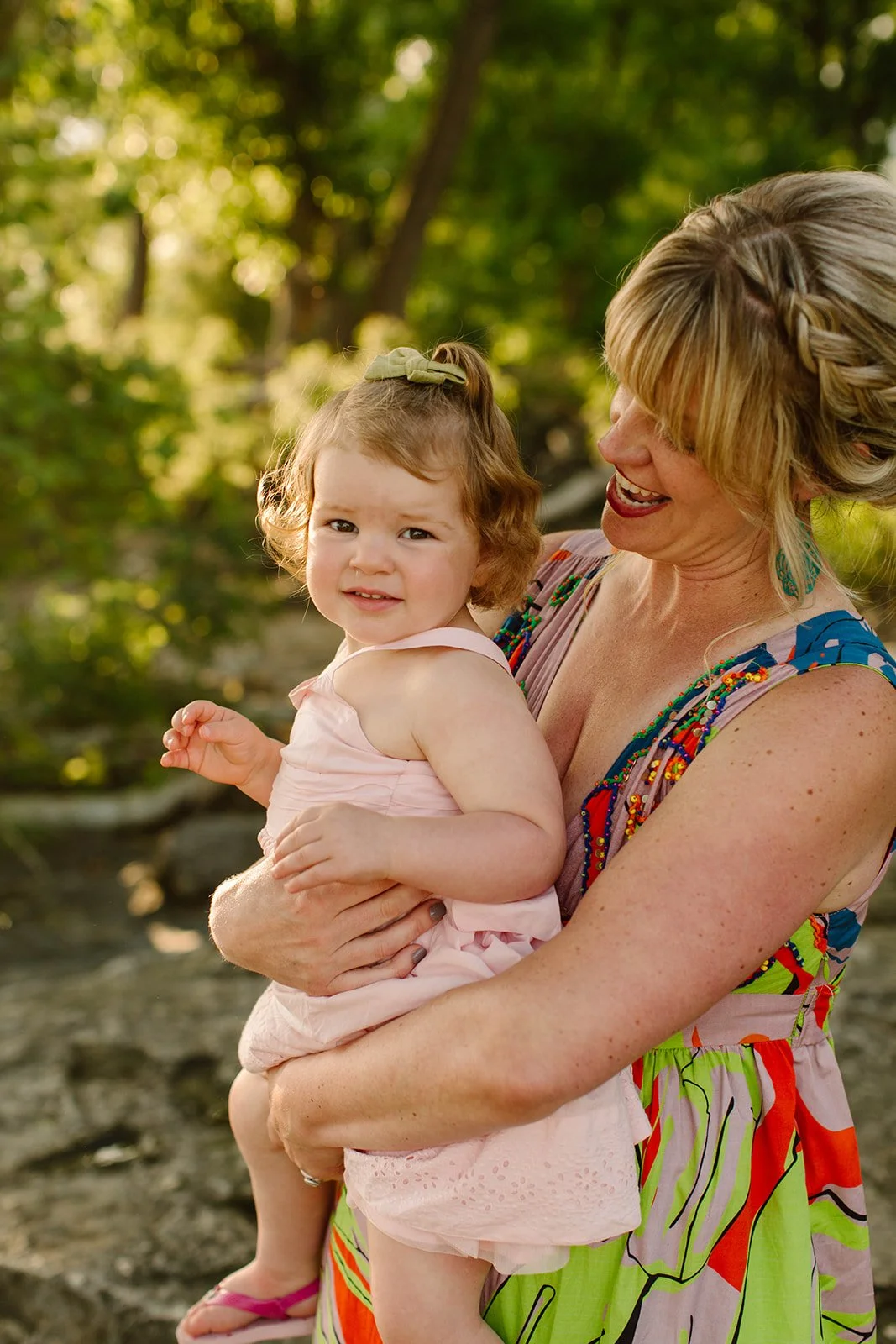 A woman smiling and holding a young girl in an outdoor setting with greenery and trees in the background.