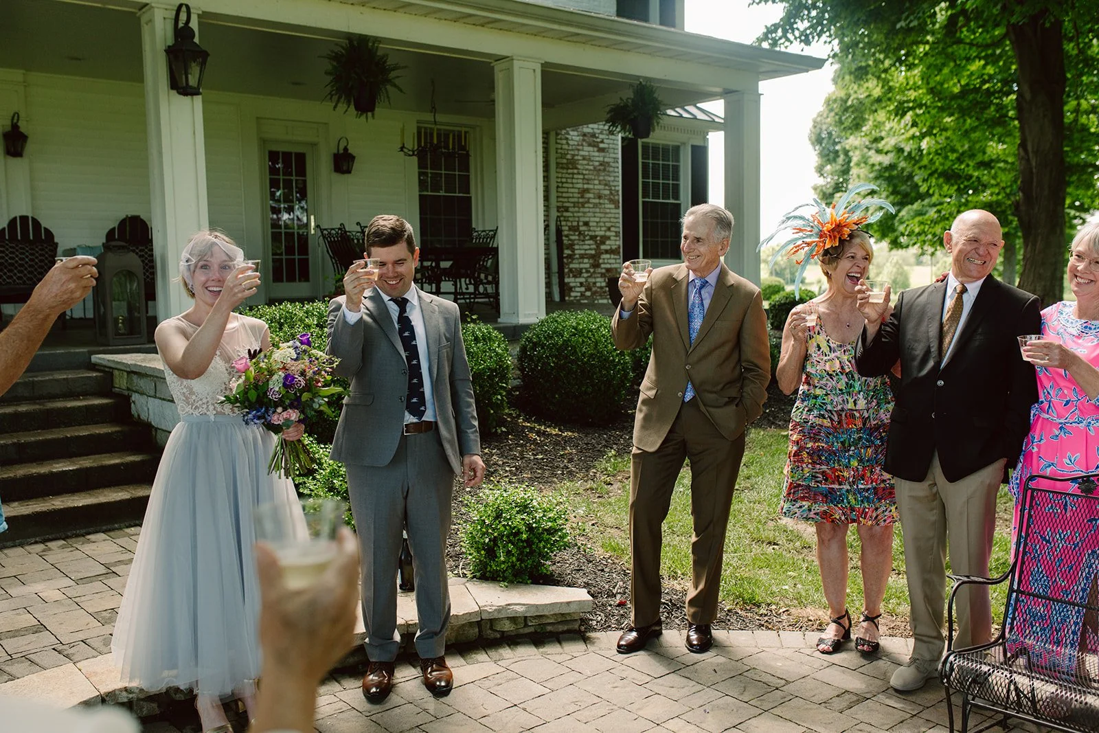 Three couples and one woman raising glasses in a toast outdoors in a garden, celebrating at a wedding reception.