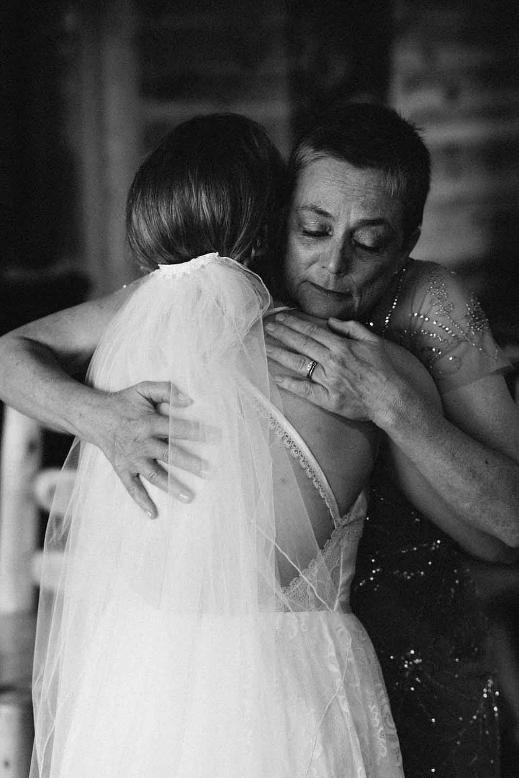 An older woman and a young girl embrace in a hug, with the woman appearing emotional. The woman is wearing a dress with embellishments, and the girl is in a wedding dress with a veil.