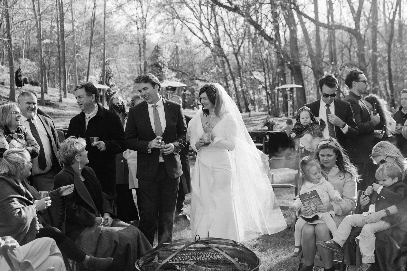 A wedding celebration outdoors with guests socializing, including a bride in a white dress and veil, surrounded by seated and standing guests, some holding drinks, in a wooded area.