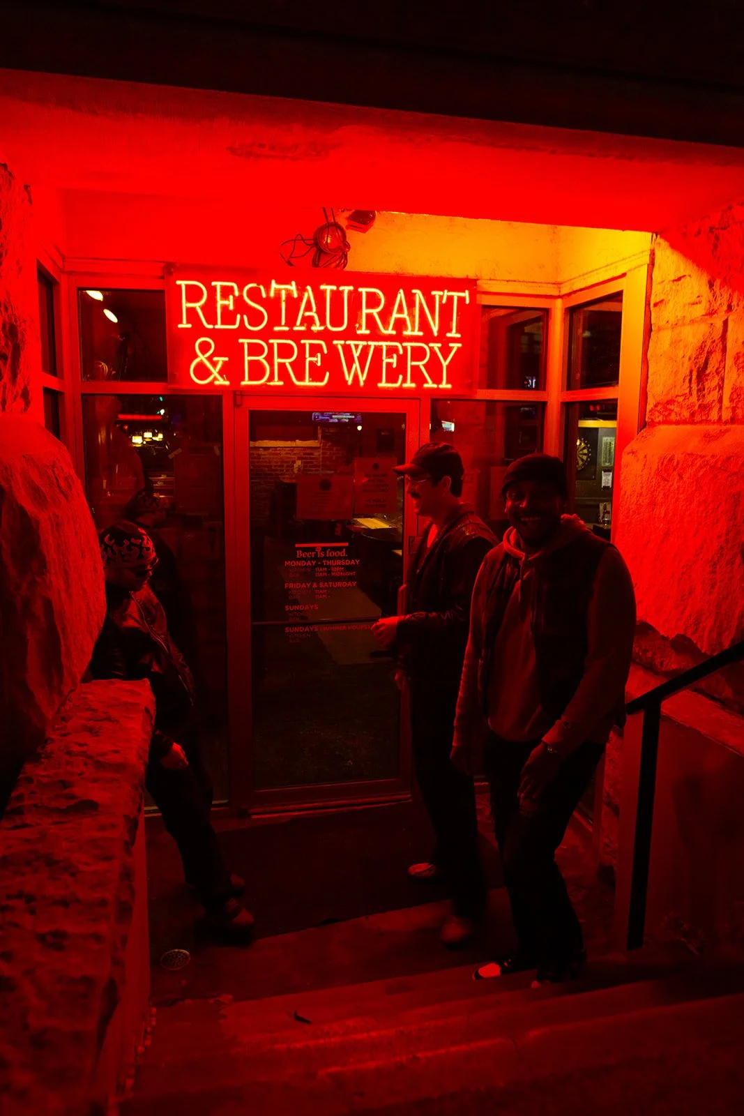 People standing under a bright red neon sign that reads 'RESTAURANT & BREWERY' outside an entrance with stone walls.