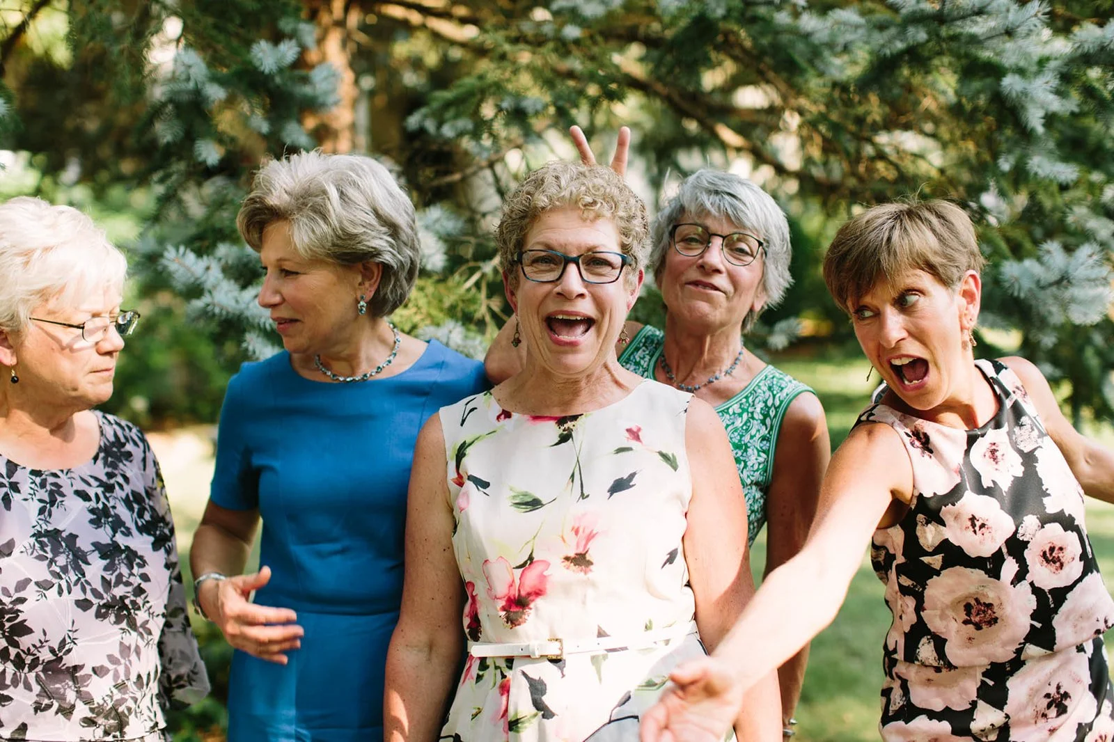 A group of five women standing outdoors in front of a large pine tree, engaging in conversation and making playful gestures.