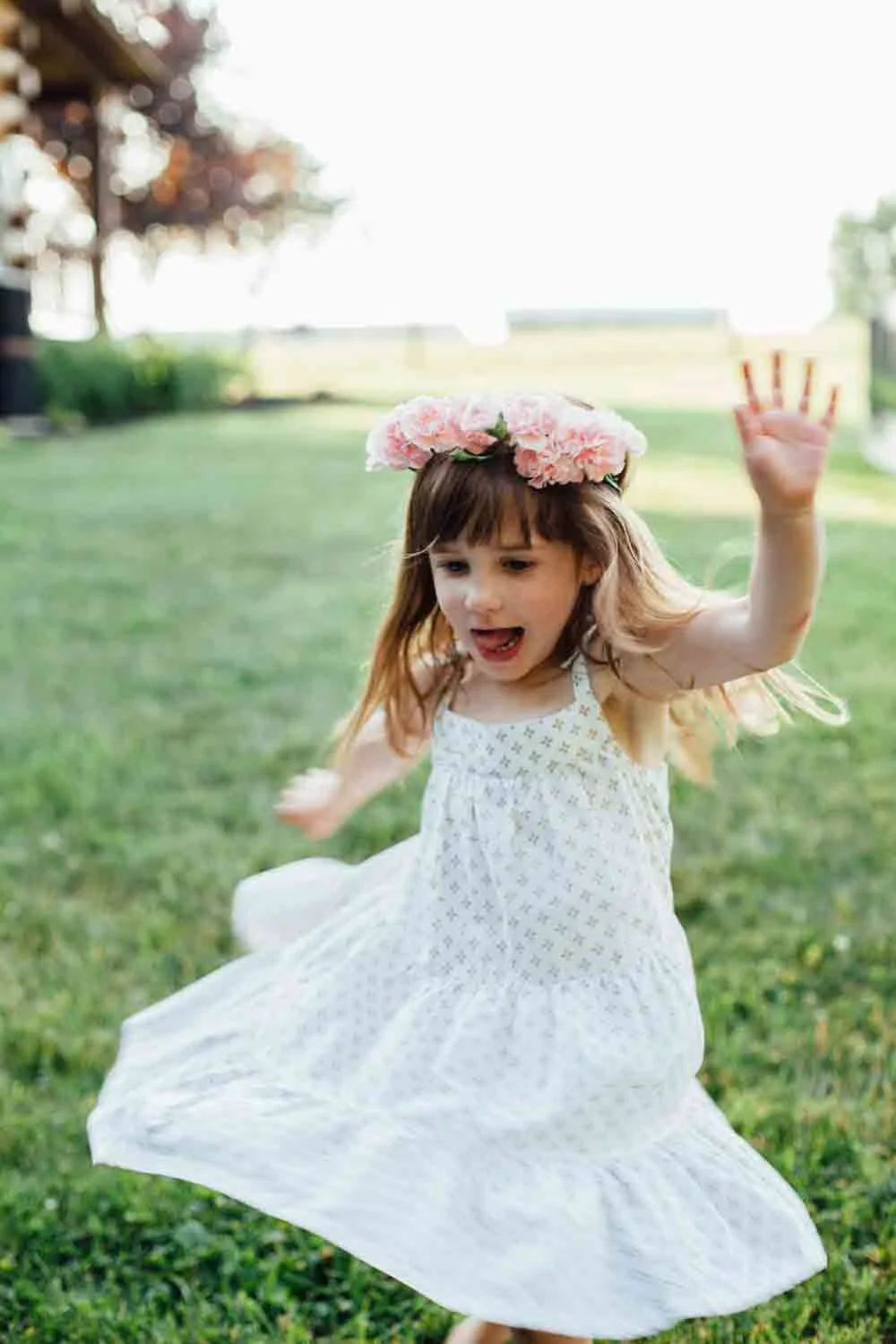 A young girl wearing a white dress and a pink flower crown is playing and jumping on a grassy area outdoors, with a blurred background of trees and a bright sky.