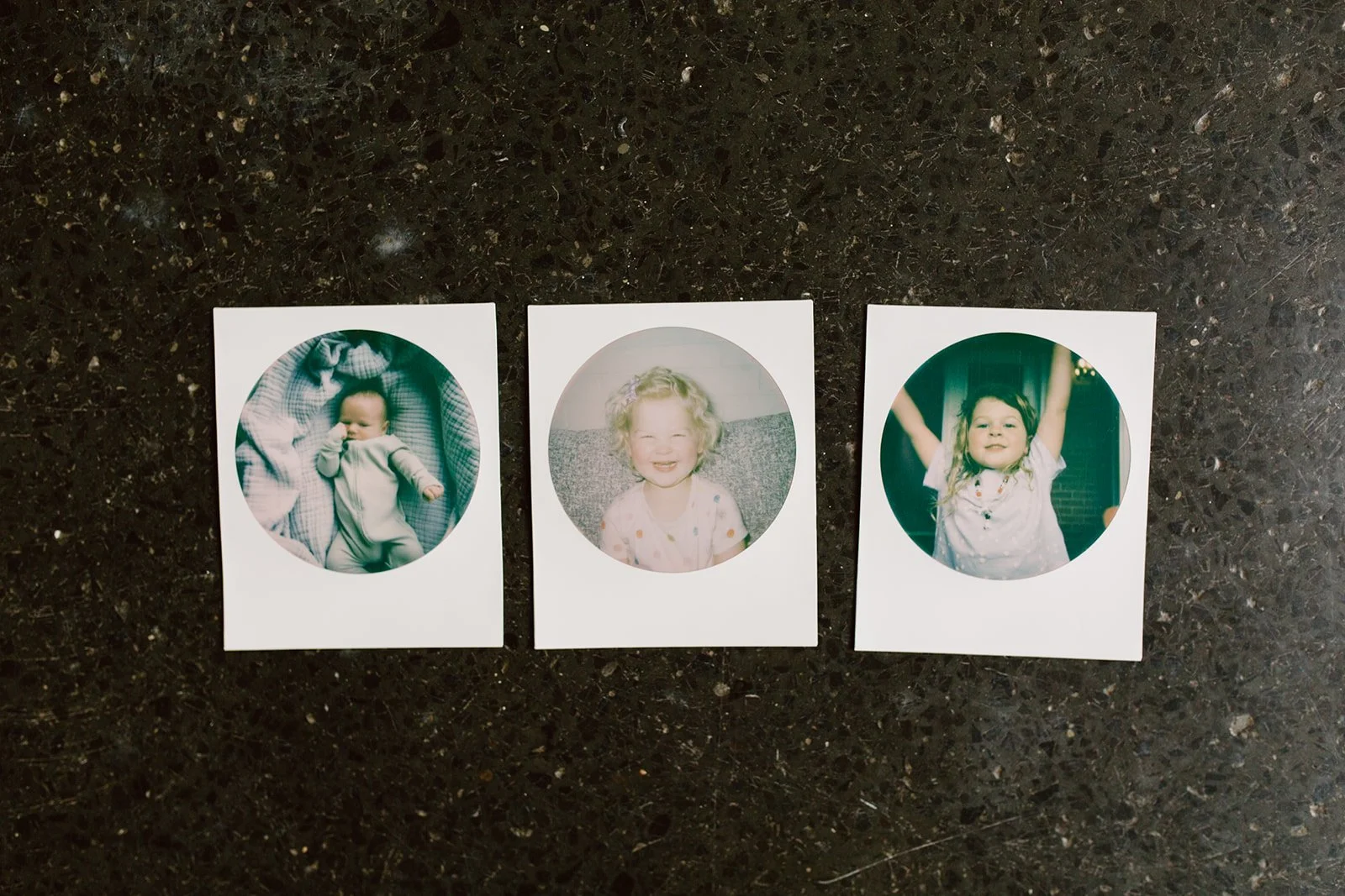 Three small photographs of young children on a dark surface, each within a white border and oval cutout. The first photo shows a baby lying down, the second a smiling girl with curly blonde hair, and the third a girl with outstretched arms and a joyful expression.