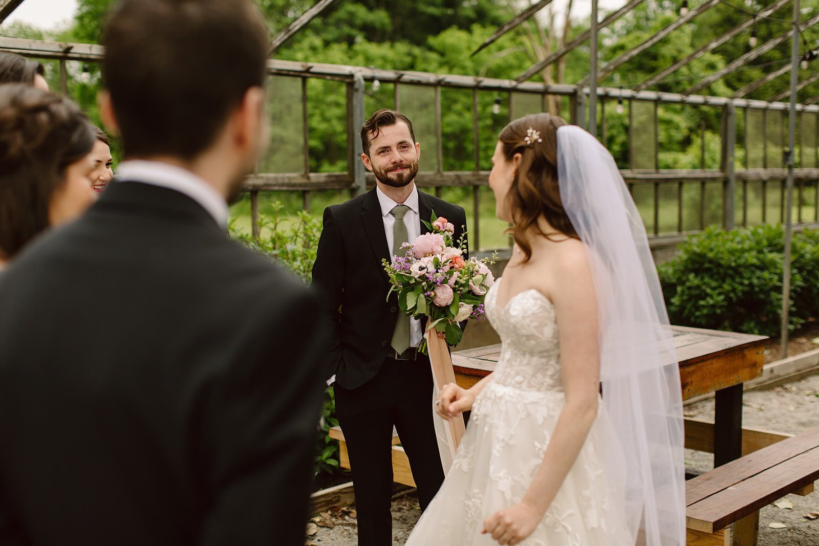 A bride and groom getting married outdoors, with the groom smiling and holding a bouquet of flowers, and the bride wearing a veil and lace dress surrounded by friends.