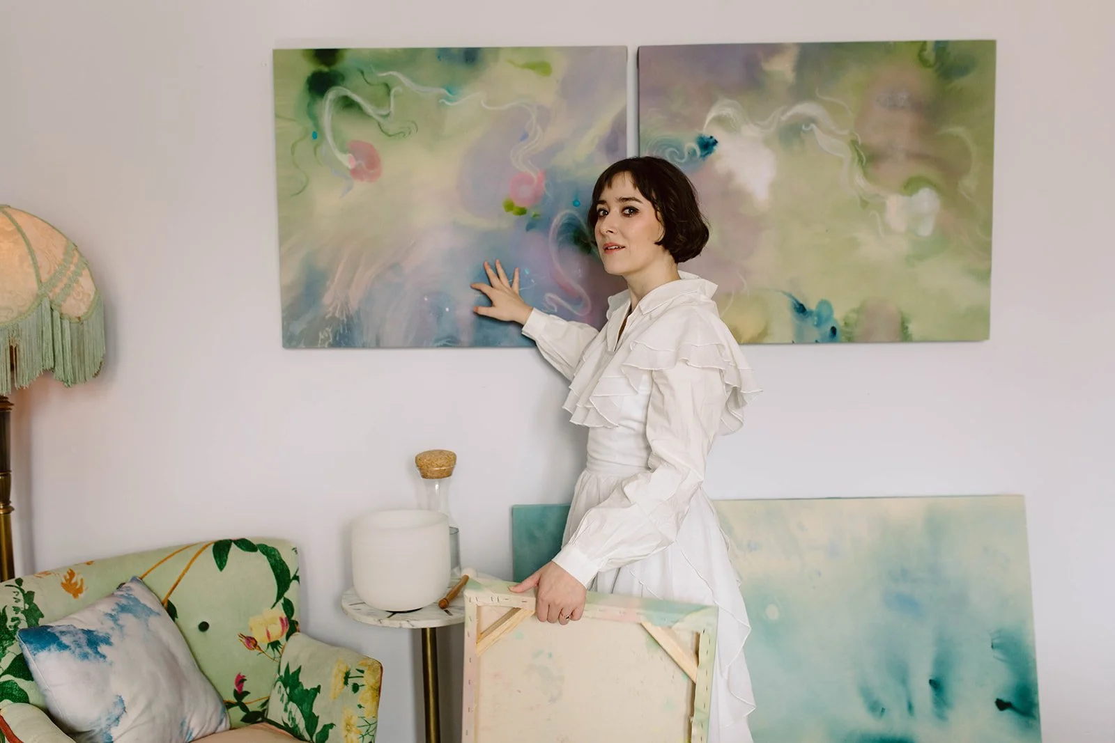 A woman with short dark hair in a white dress, standing in an art studio, touching an abstract watercolor painting on the wall. The studio has colorful artwork, a floral armchair, a lamp, and a small table with art supplies.