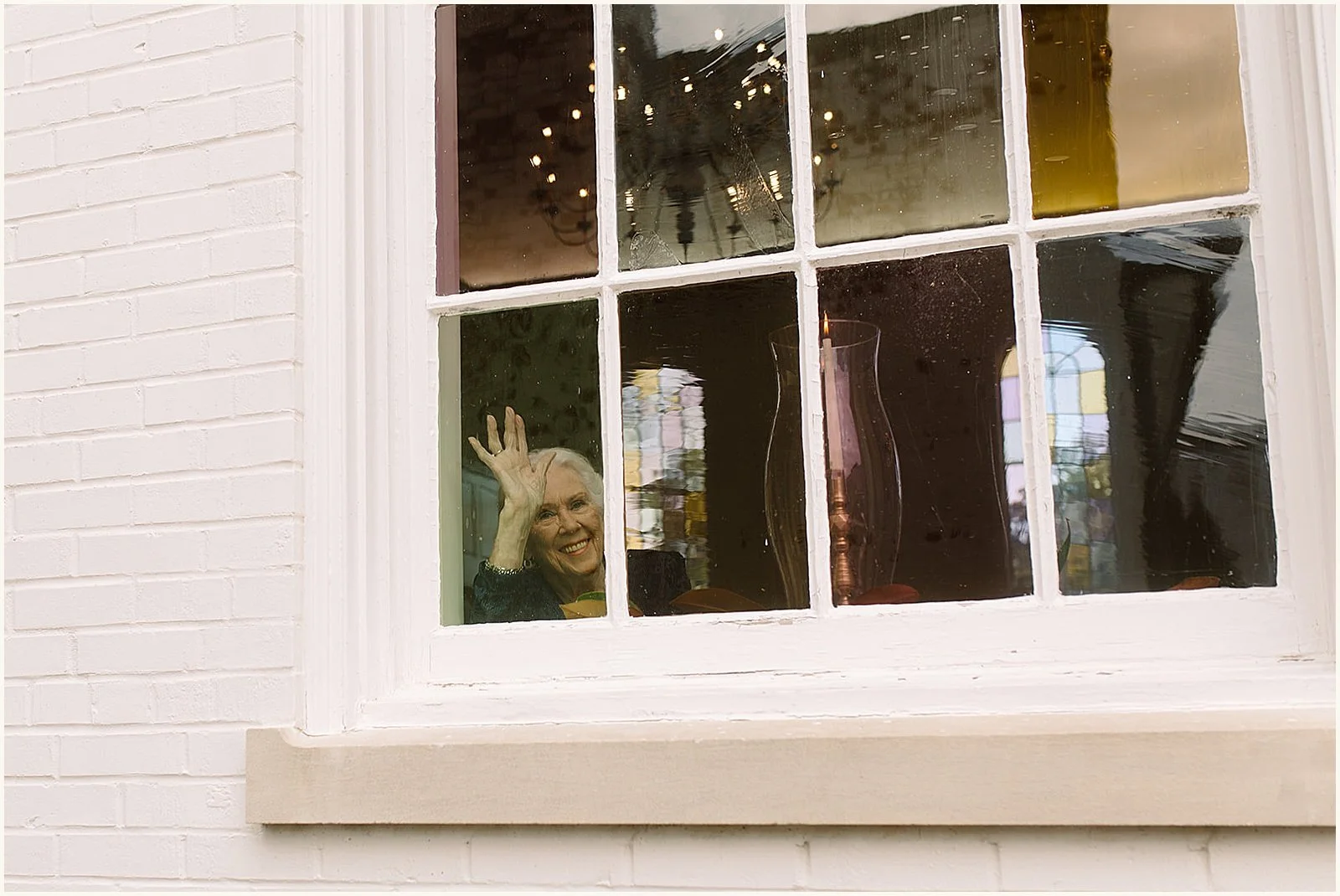 An elderly woman smiles and waves out a window.