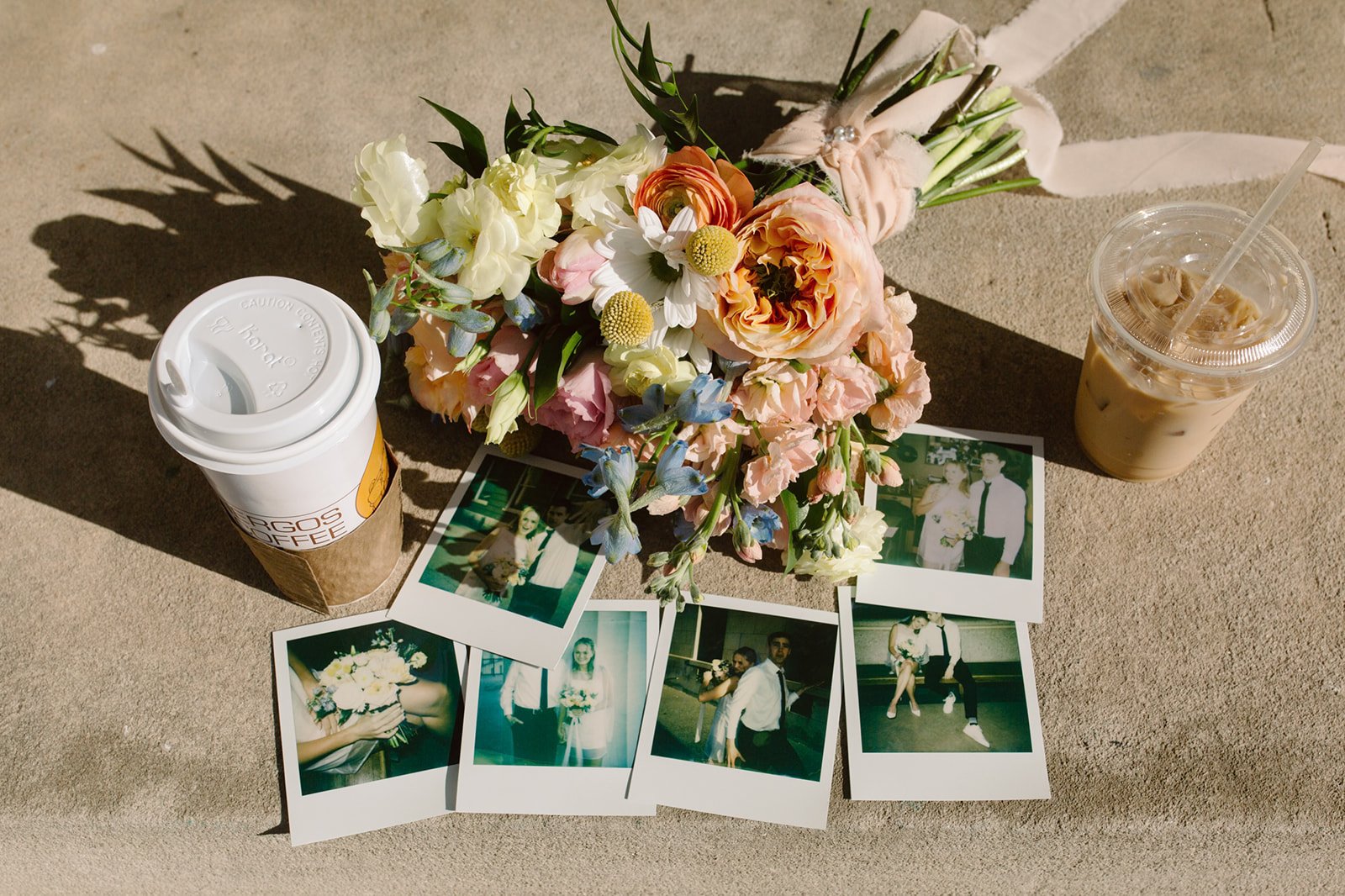 A bouquet of pink, white, and purple flowers, two iced coffee drinks, and several instant photos on a sandy surface.