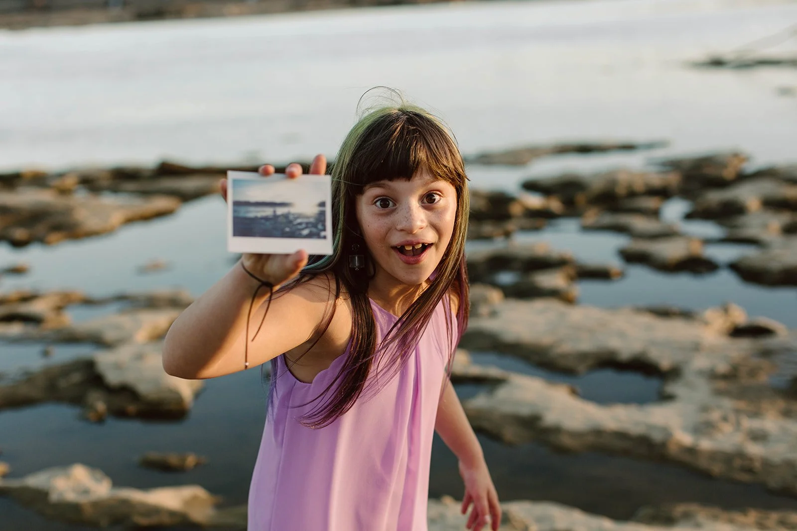 A young girl in a purple dress holding a small photograph in front of her at a rocky shoreline with water and rocks in the background.