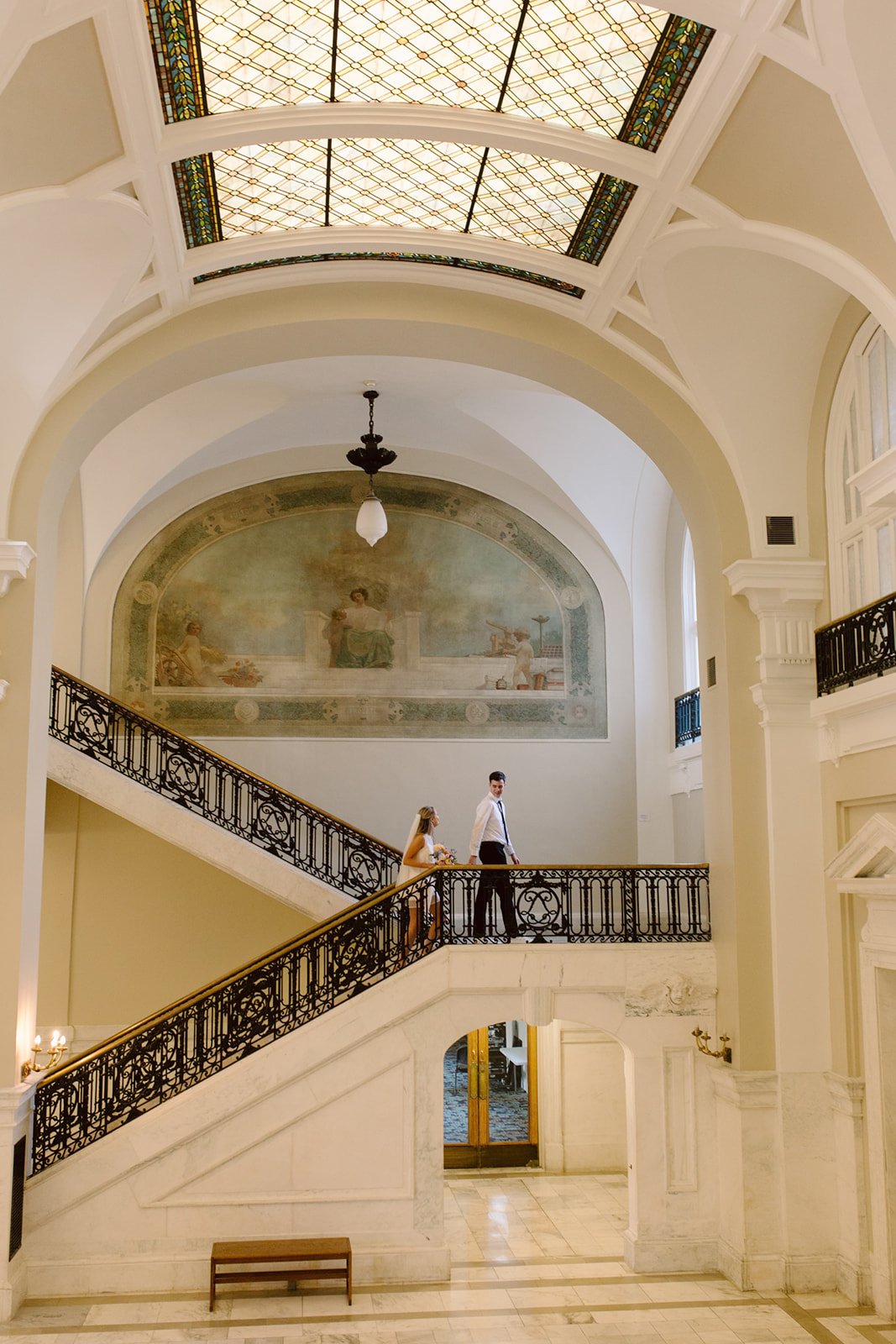 Interior view of a grand staircase in a building with ornate architectural details, including stained glass ceiling, painted mural on the wall, and two people, a man and woman, descending the staircase.