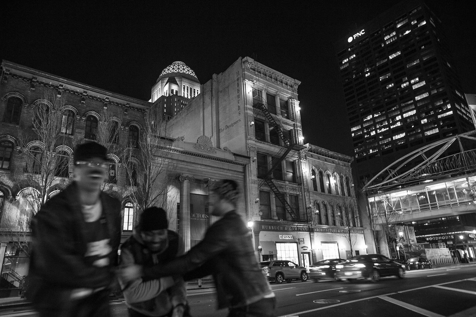 Night city street scene with tall buildings, cars, and three people in the foreground. Two are blurred and appear to be in motion, engaging in a handshake or exchange. The buildings are illuminated, with visible signage including 'PNC' on a skyscraper.