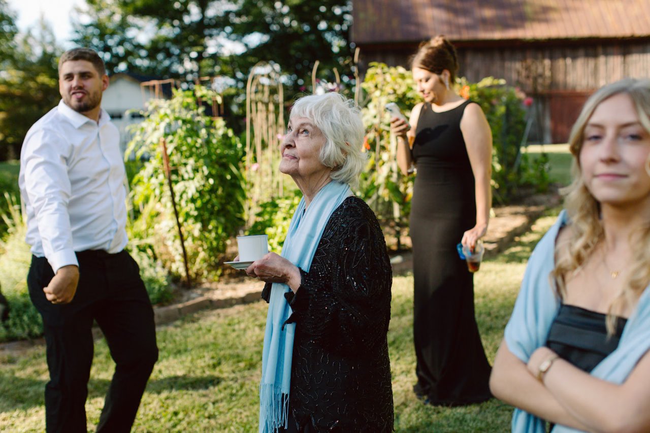 Four people at an outdoor gathering in a garden with greenery and a rustic building in the background. An elderly woman with white hair and a black dress is holding a cup and saucer. A young man in a white shirt and black pants is standing nearby. Two women are in the background, one in a black dress looking at her phone and another in a black dress with a blue shawl across her shoulders, smiling with arms crossed.