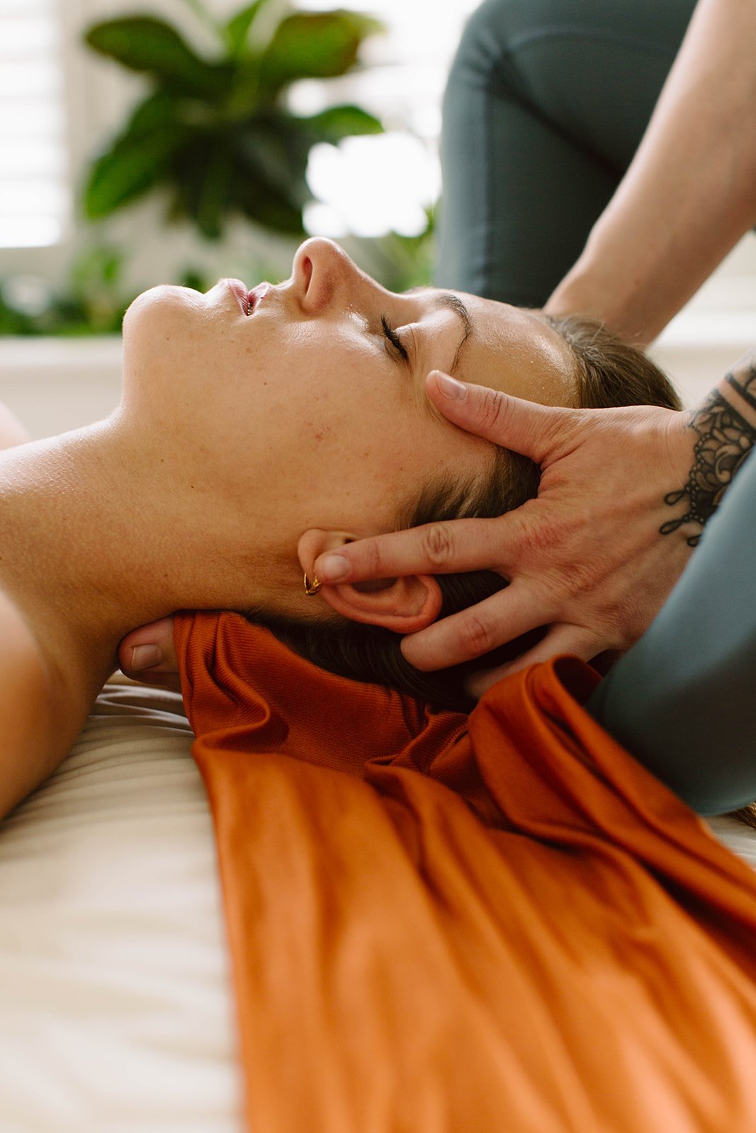 A woman receiving a neck massage, lying on a bed with her eyes closed, as a massage therapist gently supports her head.