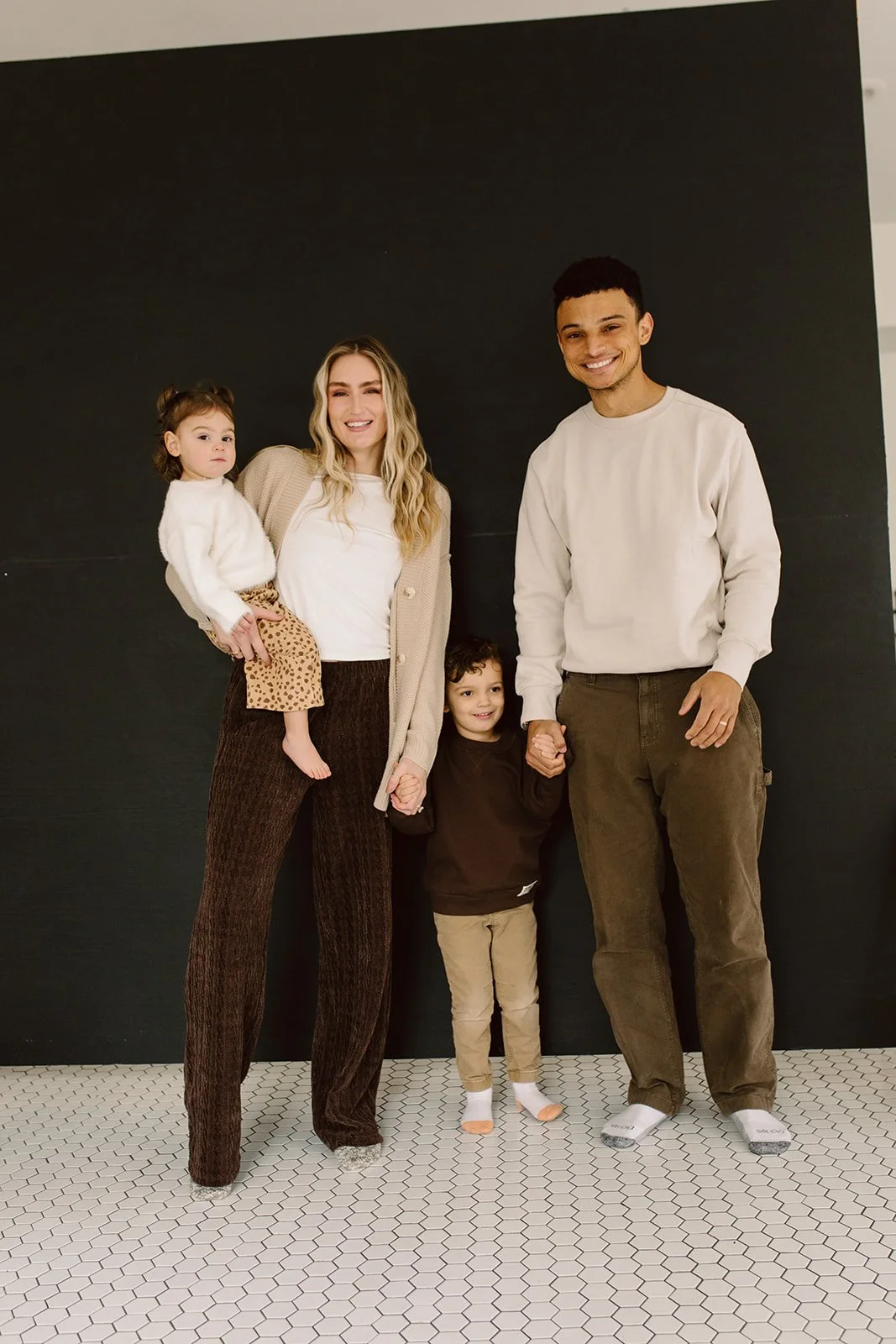 A family of four standing together, holding hands, with a black wall background and white hexagon patterned floor.
