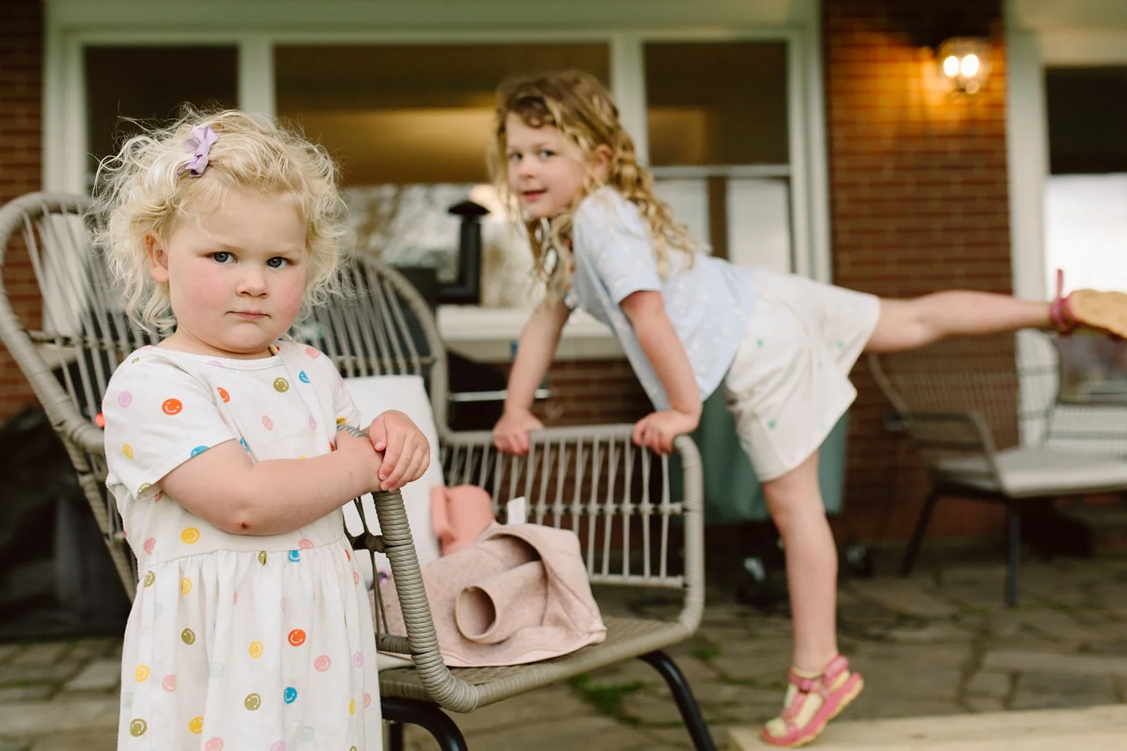 Two young girls playing outside on a patio. One girl in the foreground wears a white dress with colorful smiley faces, looking serious. The other girl in the background, wearing a light-colored shirt and shorts, is climbing on patio furniture.