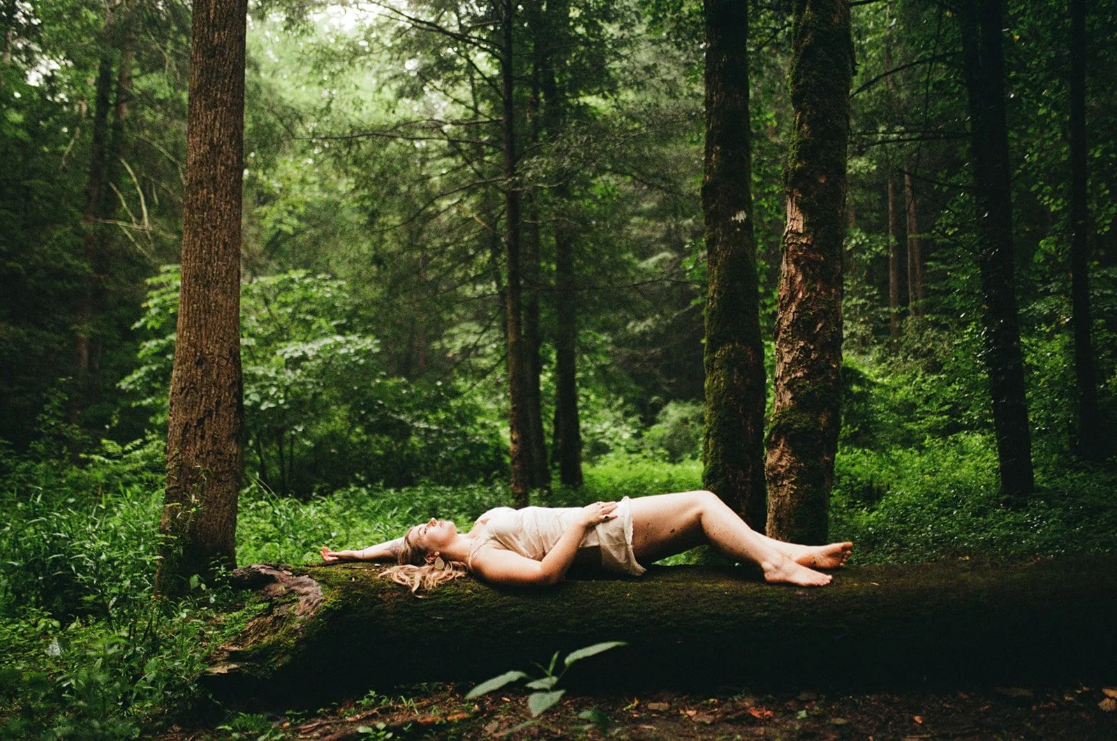 A woman lying on her back on a fallen tree trunk in a lush green forest, with her arms stretched above her head and eyes closed.