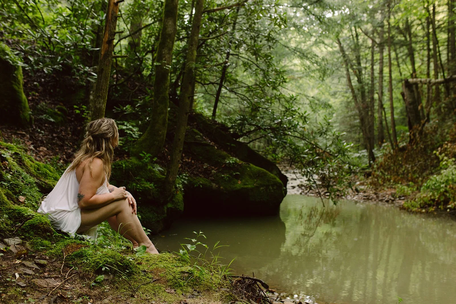 A woman sitting on mossy ground at the edge of a creek in a dense, green forest.