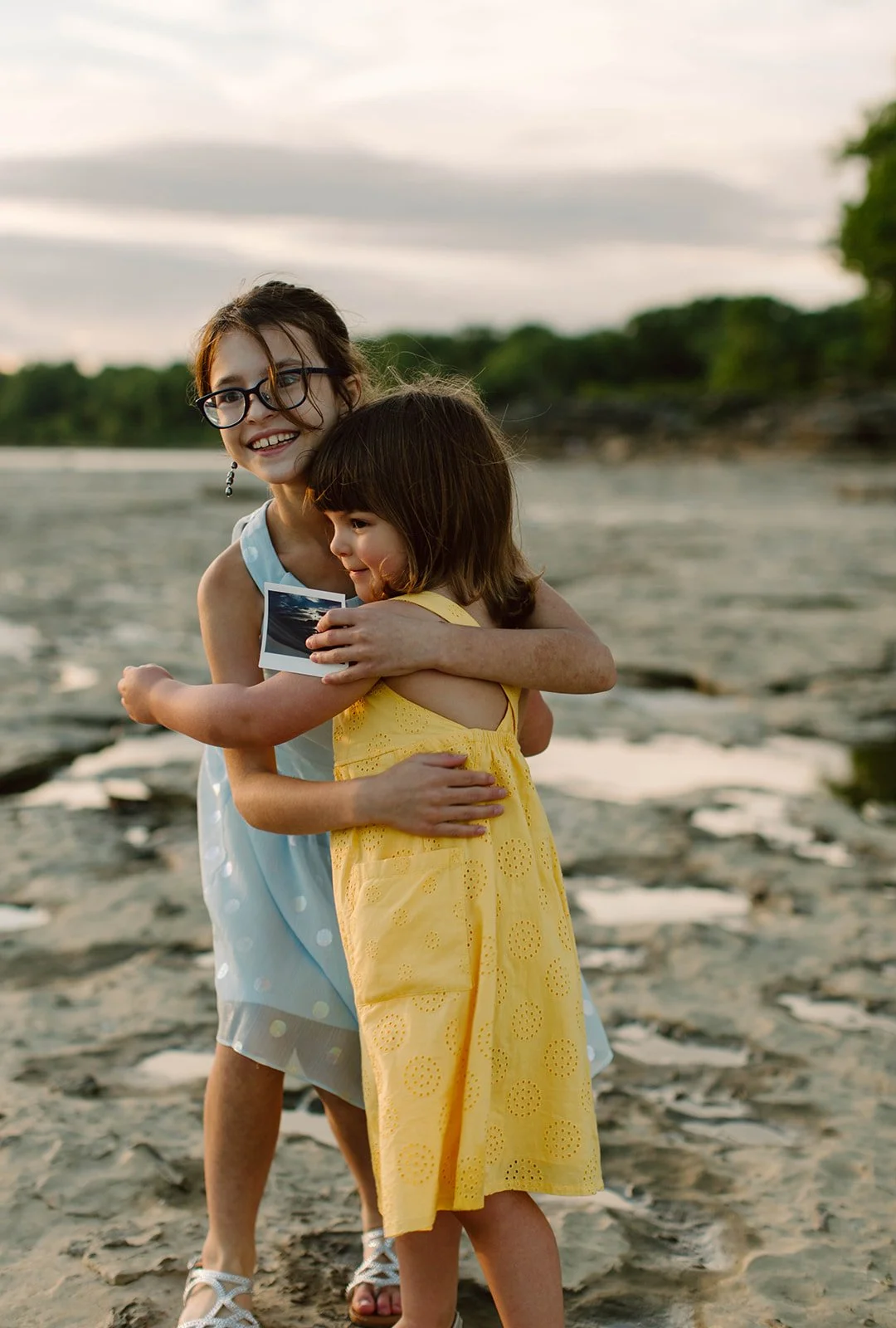 Two young girls hugging on a sandy beach at sunset, one holds an ultrasound photo.