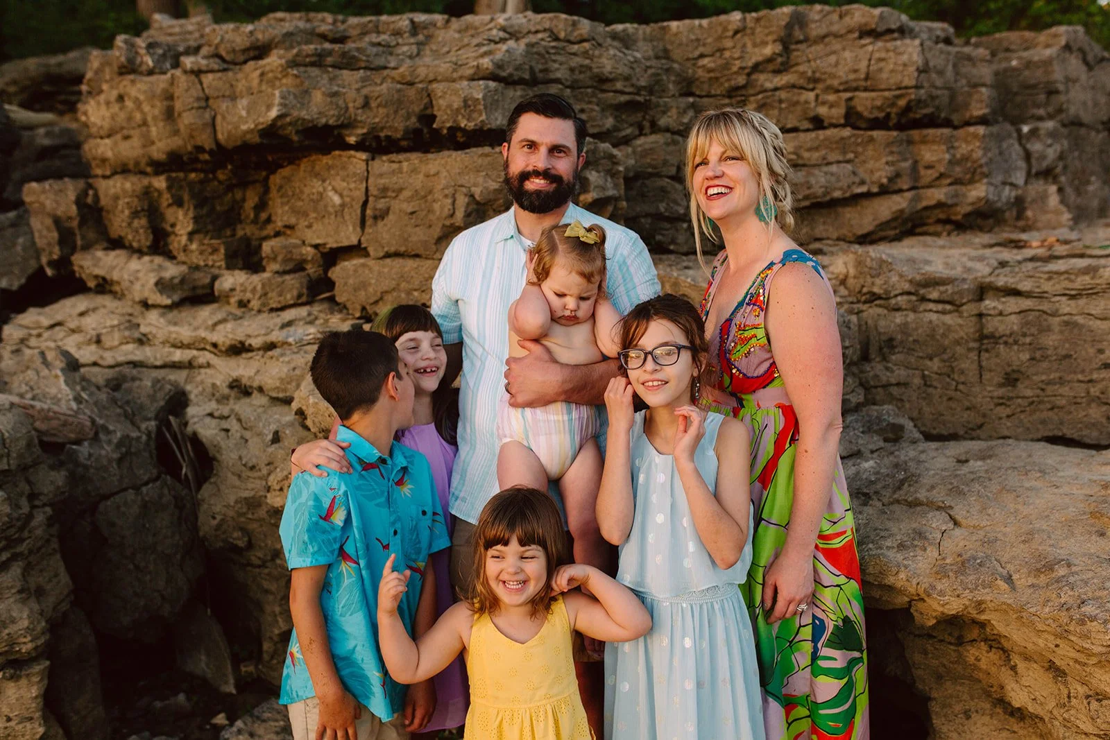 A family of seven standing outdoors against a rocky background during sunset, smiling and laughing, with children wearing colorful summer clothing.